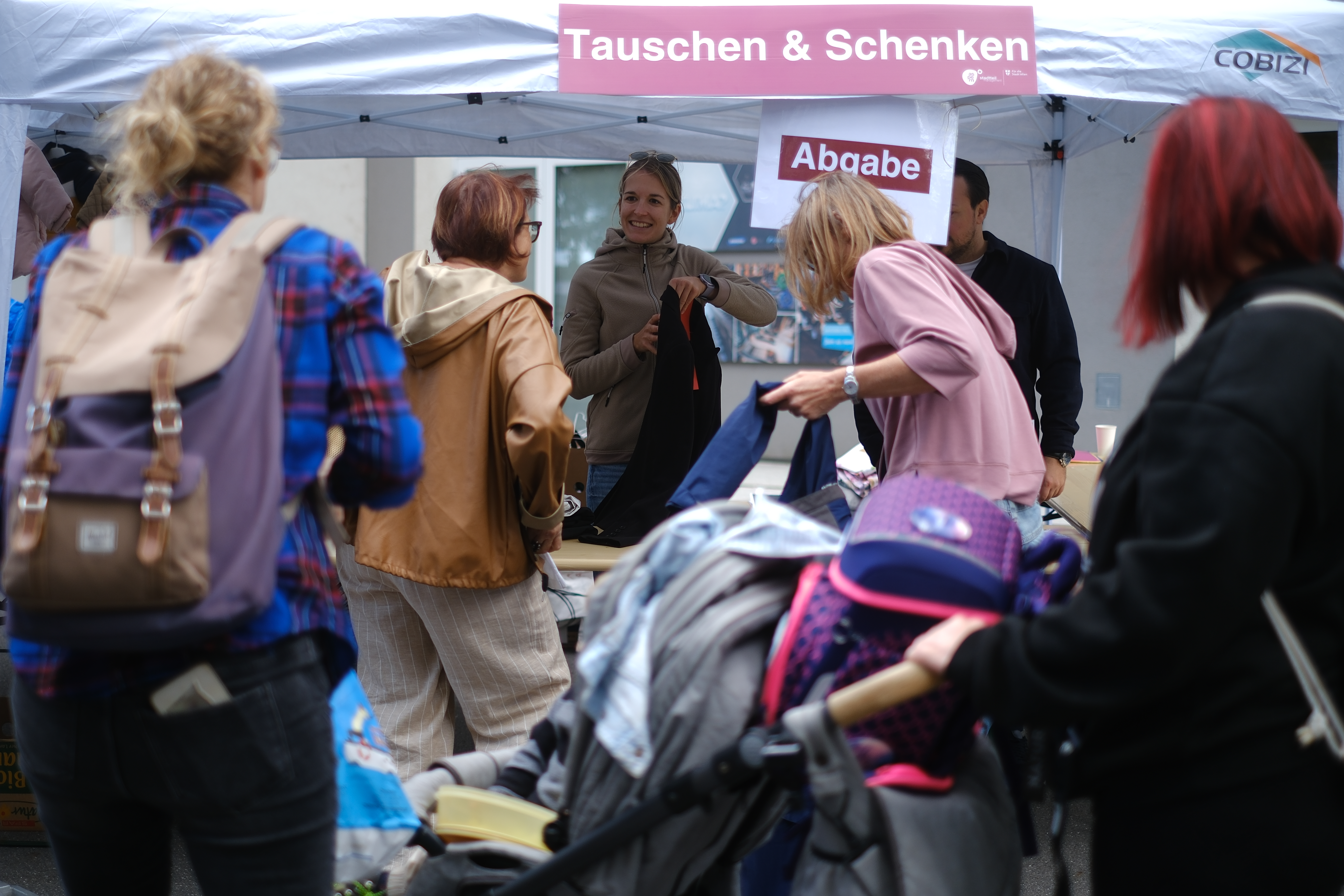 Personen stehen vor einem Pavillon mit der Aufschrift Tauschen & Schenken und geben Kleidung ab