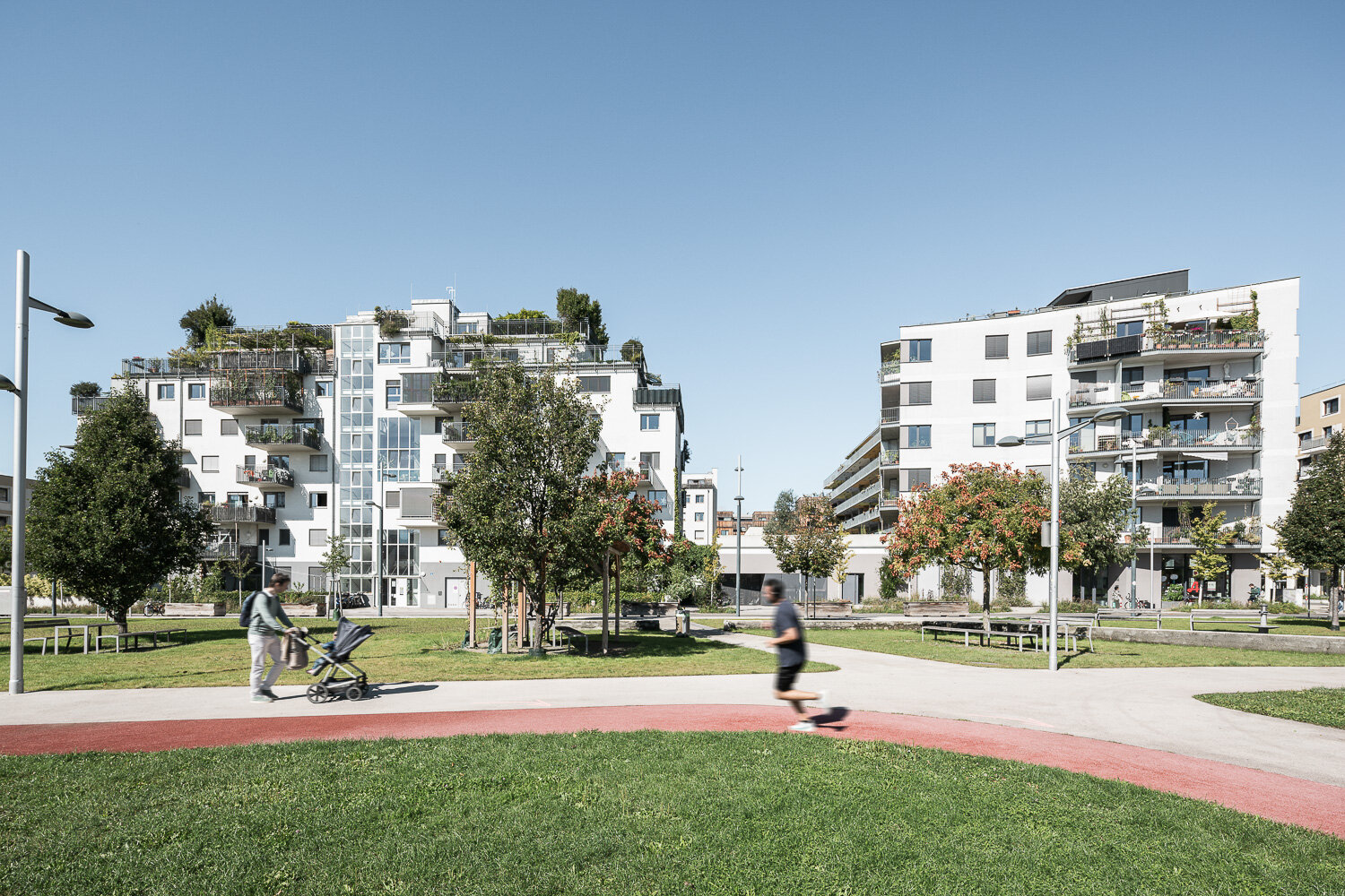 Moderne Wohngebäude mit Parkanlage und Spaziergängern Zwei moderne Wohngebäude mit Balkonen und Begrünung, davor Parkanlage mit Spaziergängern und Bäumen.