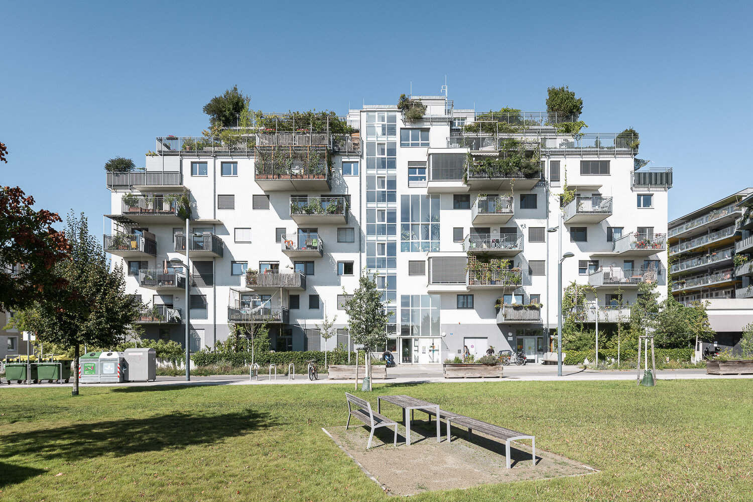 Begrünte Wohnanlage mit Balkonen und Dachgärten Modernes, mehrstöckiges Wohngebäude mit bepflanzten Balkonen und Dachgärten, umgeben von Rasenflächen und Bäumen bei klarem, blauem Himmel.