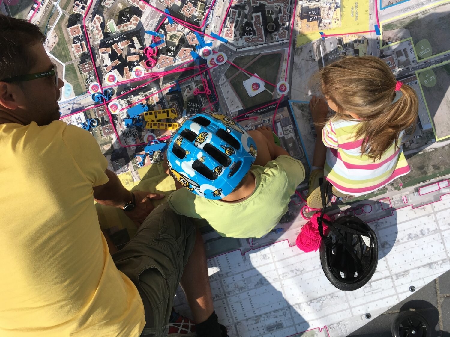 Matte mit der Seestadt abgebildet, darauf ein Mann und zwei Kinder, eines mit Helm, die sich die Matte ansehen.