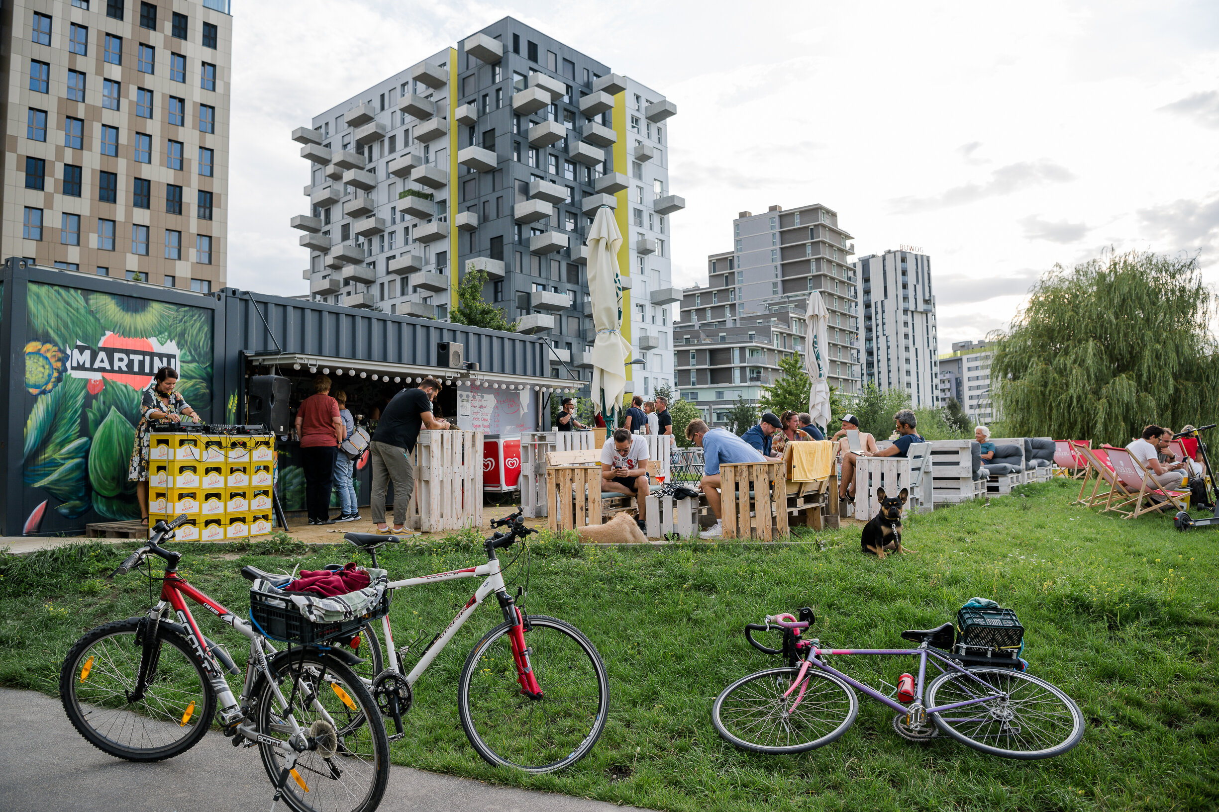 Fahrräder in Wiese und Menschen vor einem bunt bemalten Bar-Container im Park vor modernen Häusern