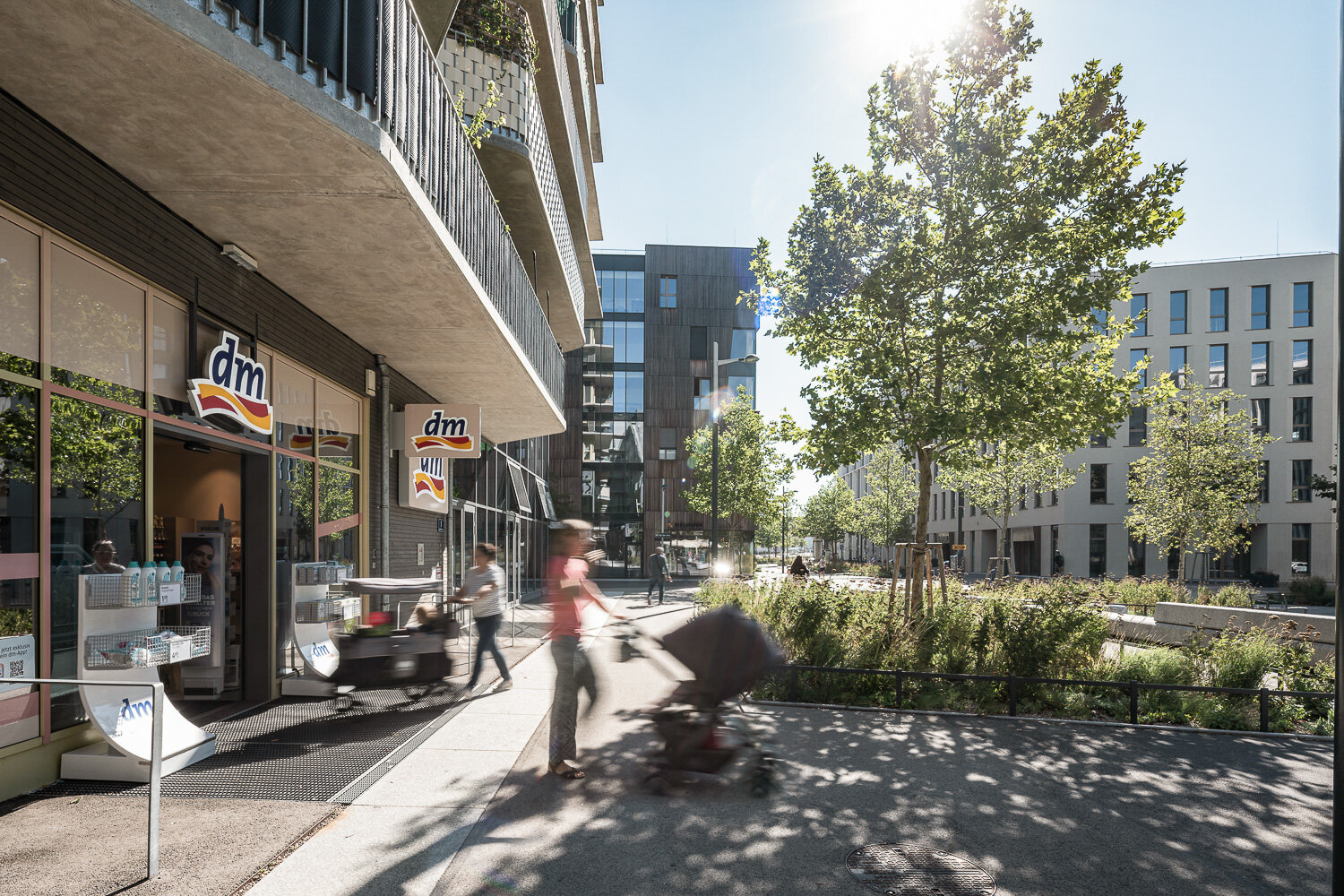 geräumiger strak begrünter Platz zwischen Wohn- und Bürogebäuden und Menschen vor einem Drogeriemarkt