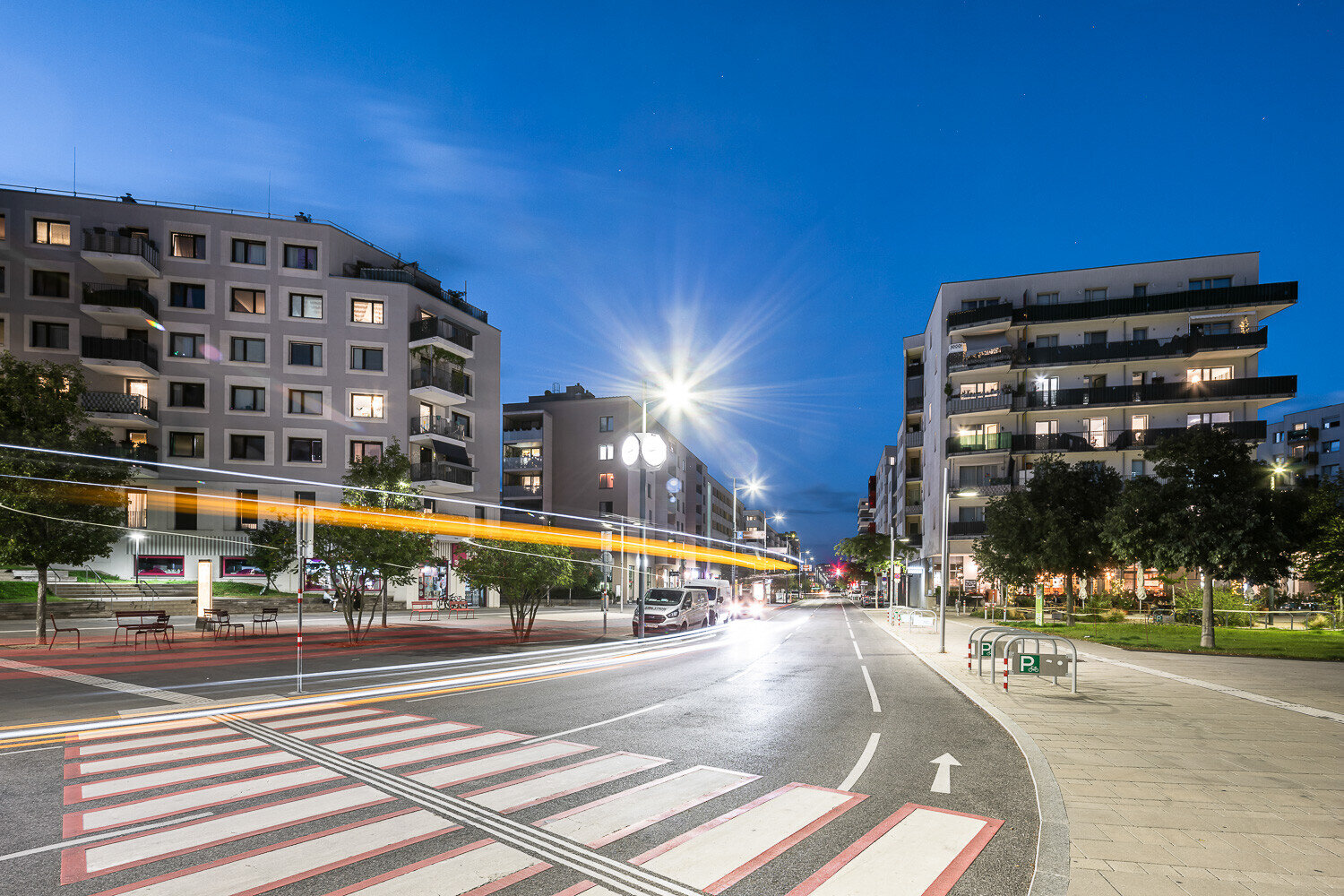 Abendstimmung in der Seestadt: moderne Wohngebäude, beleuchtete Straßenlaternen und Lichtspuren eines vorbeifahrenden Busses auf der zentralen Straße, mit Zebrastreifen und Radweg im Vordergrund.