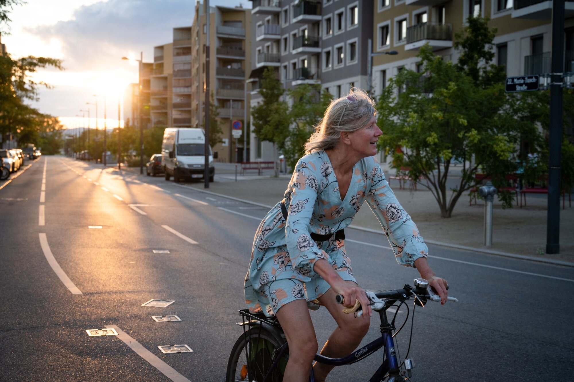 ältere Frau mit Sommerkleid fährt entspannt in einer Allee mit modernen Wohnhäusern mit dem Rad im Sonnenuntergang