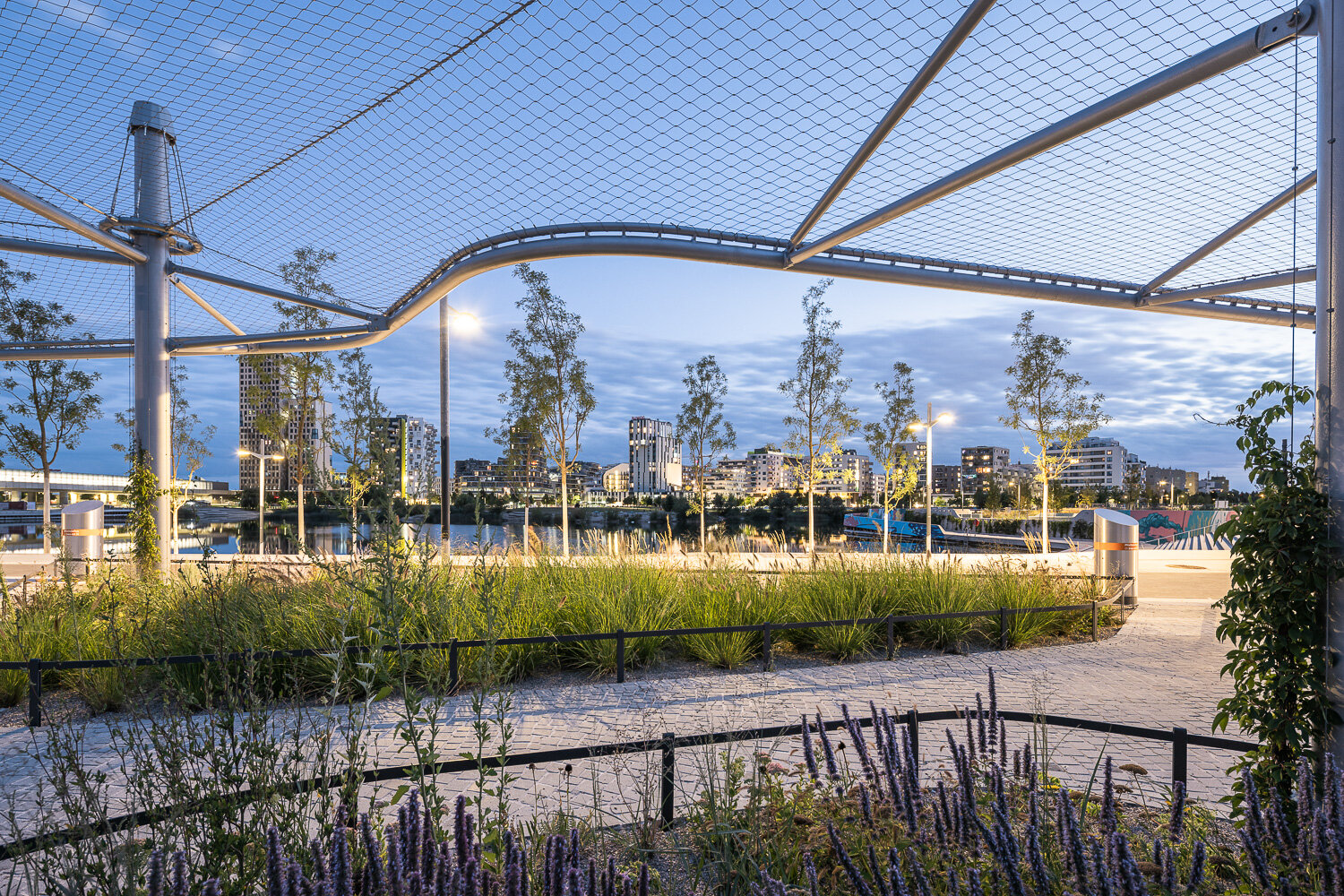 Beete, Bäume und eine offene Überdachung mit Wegen vor der Skyline der Seestadt