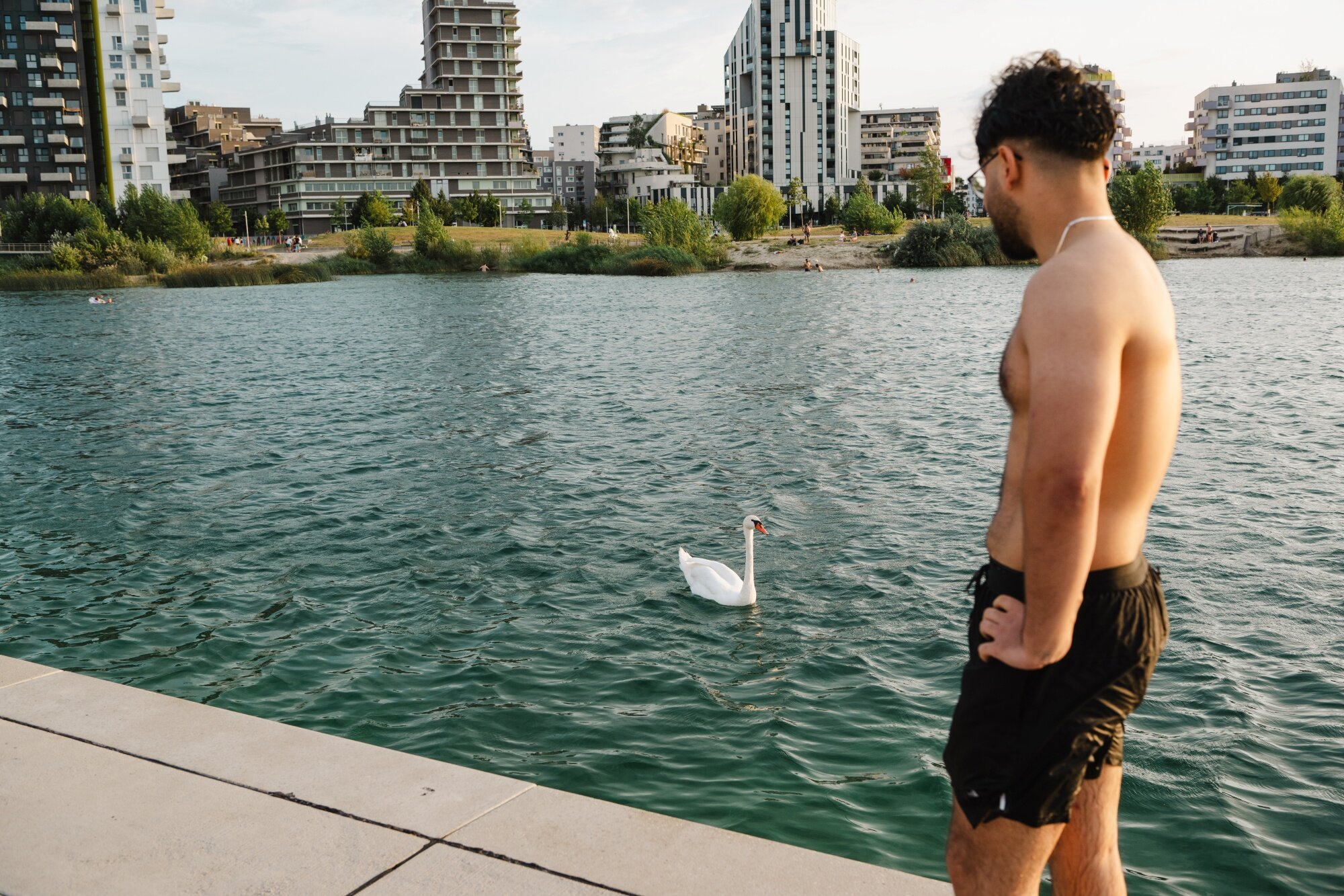 Urlaubsgefühl am See in der Seestadt Mann mit Badehose betrachtet Schwan im blauen See mit urbaner Skyline und grünen Uferanlagen