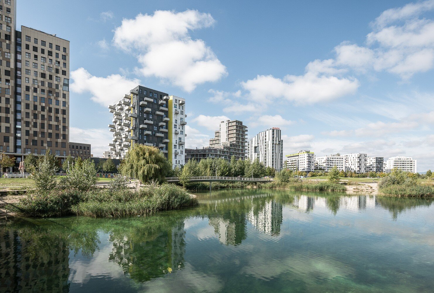 See mit blaugrünem Wasser und begrünten Wildbadezugängen vor modernen Hochhäusern und Wohnbauten