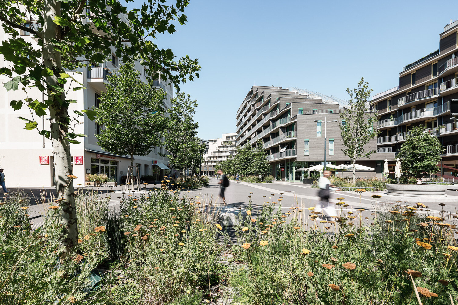 Urbanes Grün und kurze Wege Menschen auf einem großen Platz mit viel Begrünung, Lokalen und Geschäften sowie modernen Gebäuden
