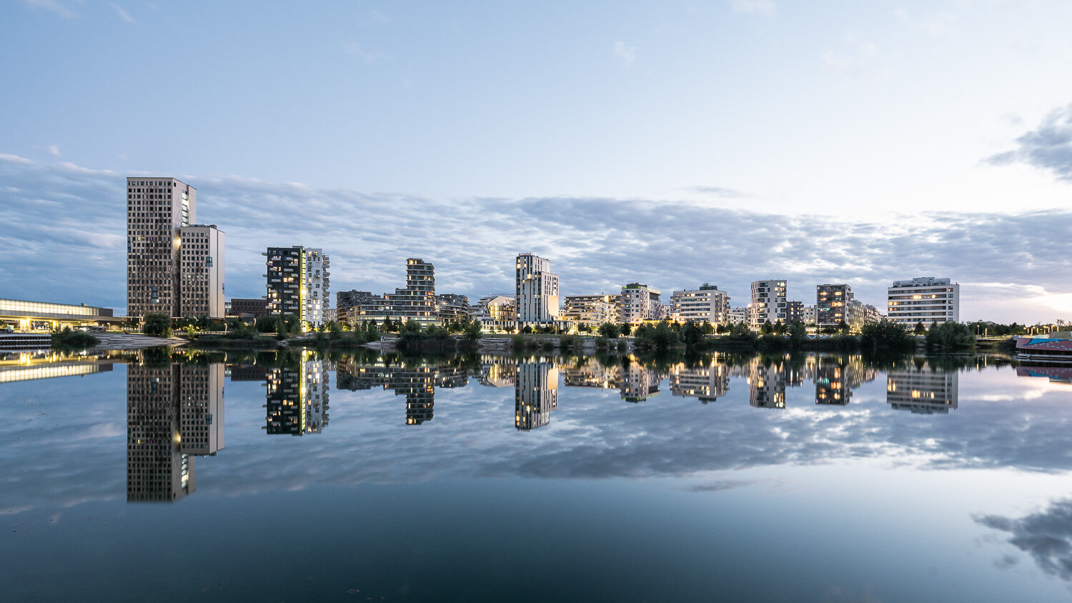 hochmoderne Skyline eines Wirtschaftsstandorts mit Bürogebäuden und Wohngebäuden hinter einem See mit intensiv begrüntem Ufer