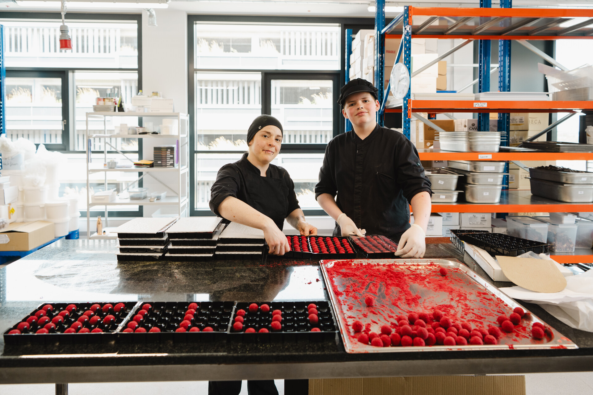 Xocolat verbindet Genuss mit nachhaltiger Produktion - inmitten der Seestadt. Zwei Personen arbeiten an der Pralinenproduktion. Die Pralinen sind rot und formen das Wort "Hello".