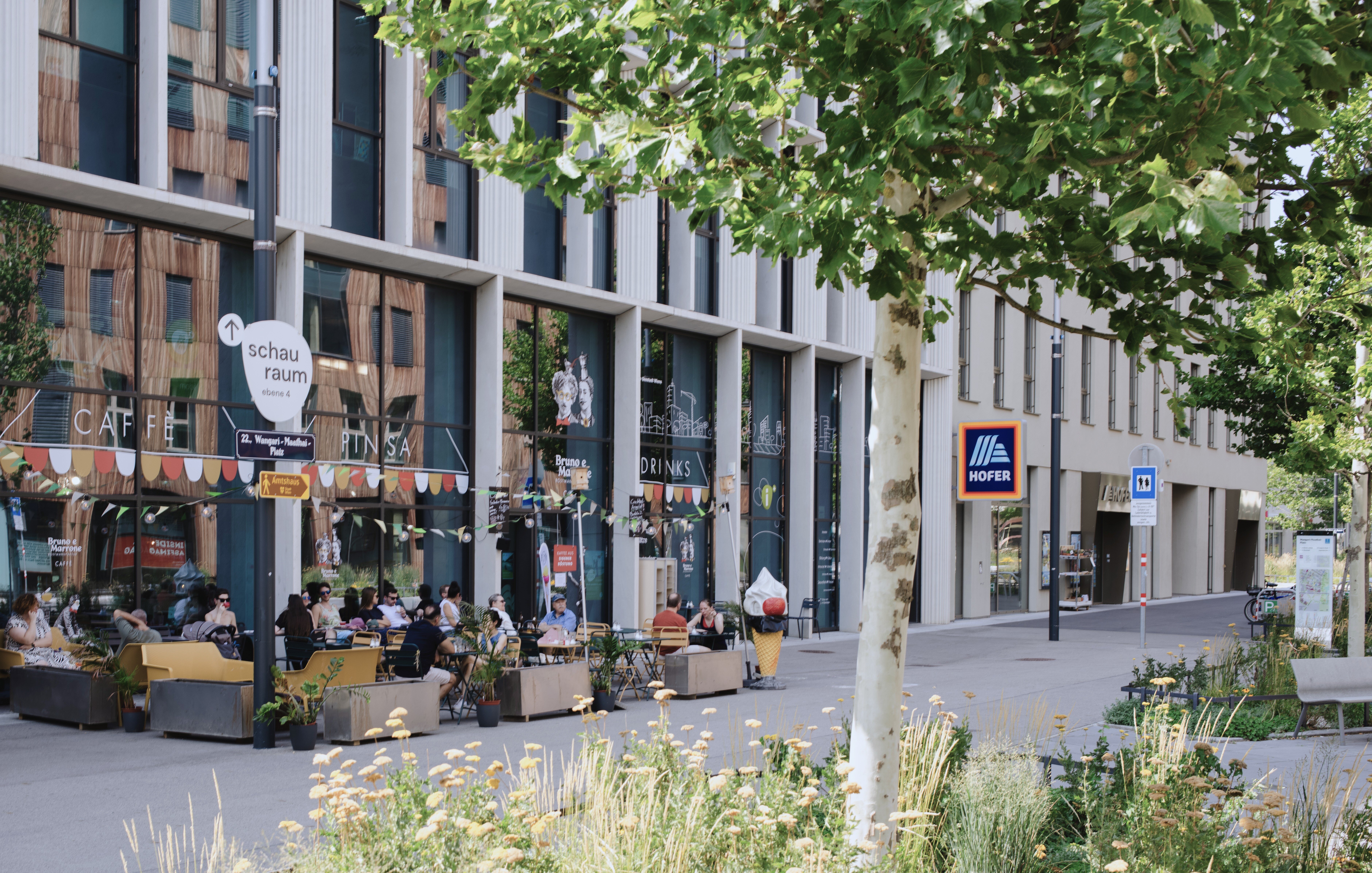 viele Menschen in einen Gastgarten eines hippen Lokales auf einem begrünten Platz mit weiteren Geschäften