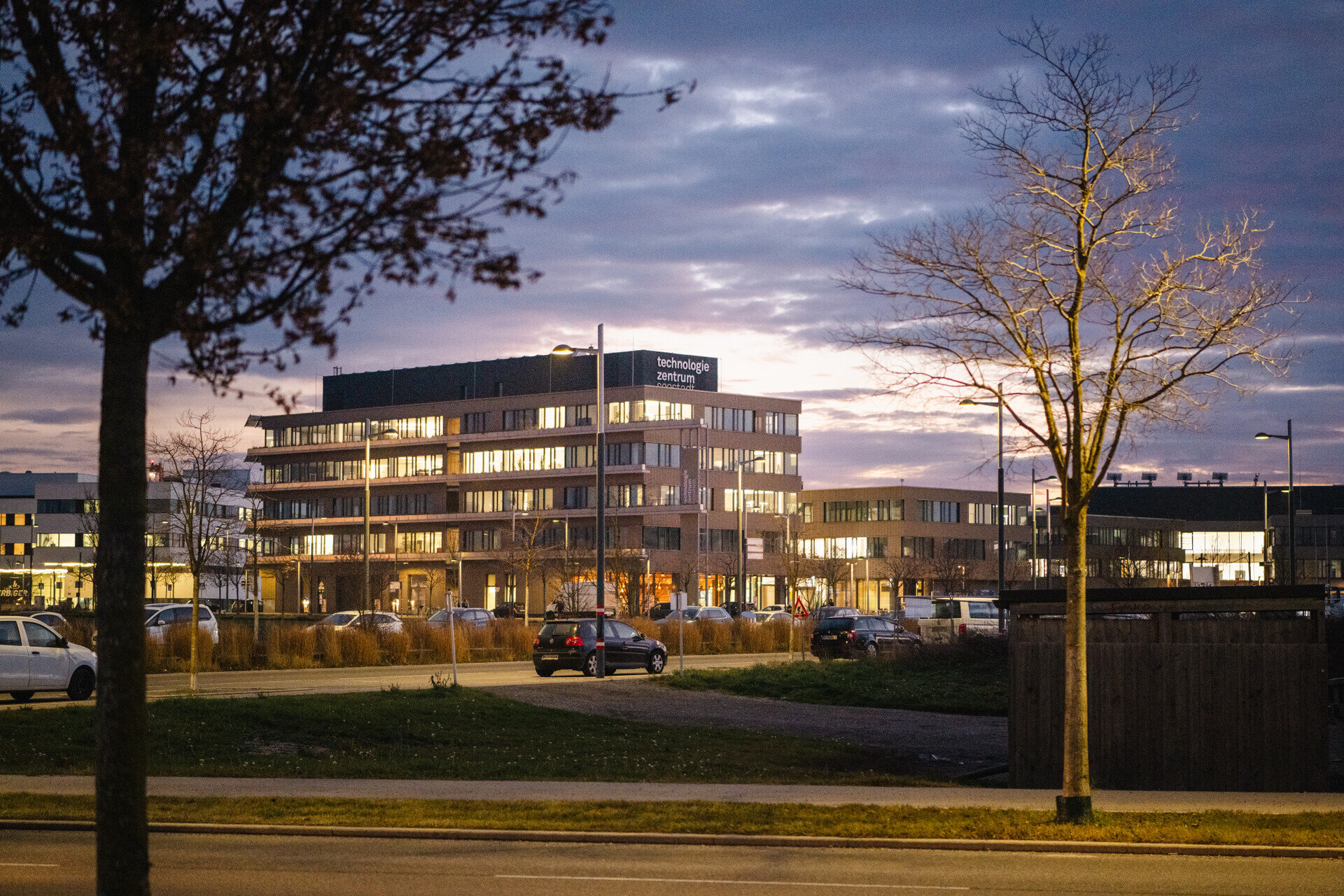 Abendaufnahme des Technologiezentrums Seestadt mit beleuchteten Büros, modernen Gebäuden und parkenden Autos im Vordergrund.