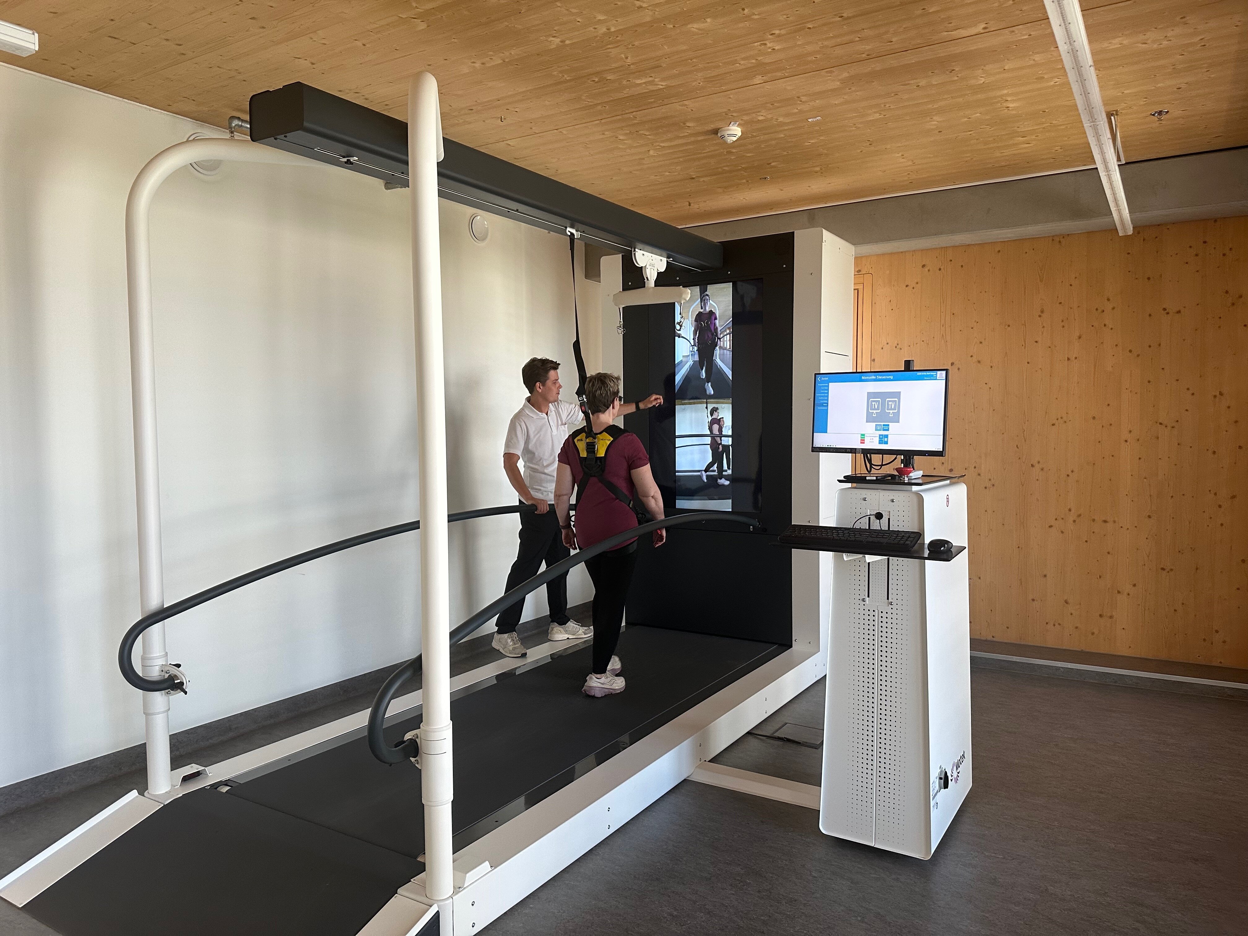 Eine Frau steht auf einem Laufband, vor ihr ist ein Bildschirm zu sehen, der Therapeut zeigt auf den Bildschirm und erklärt die Übung