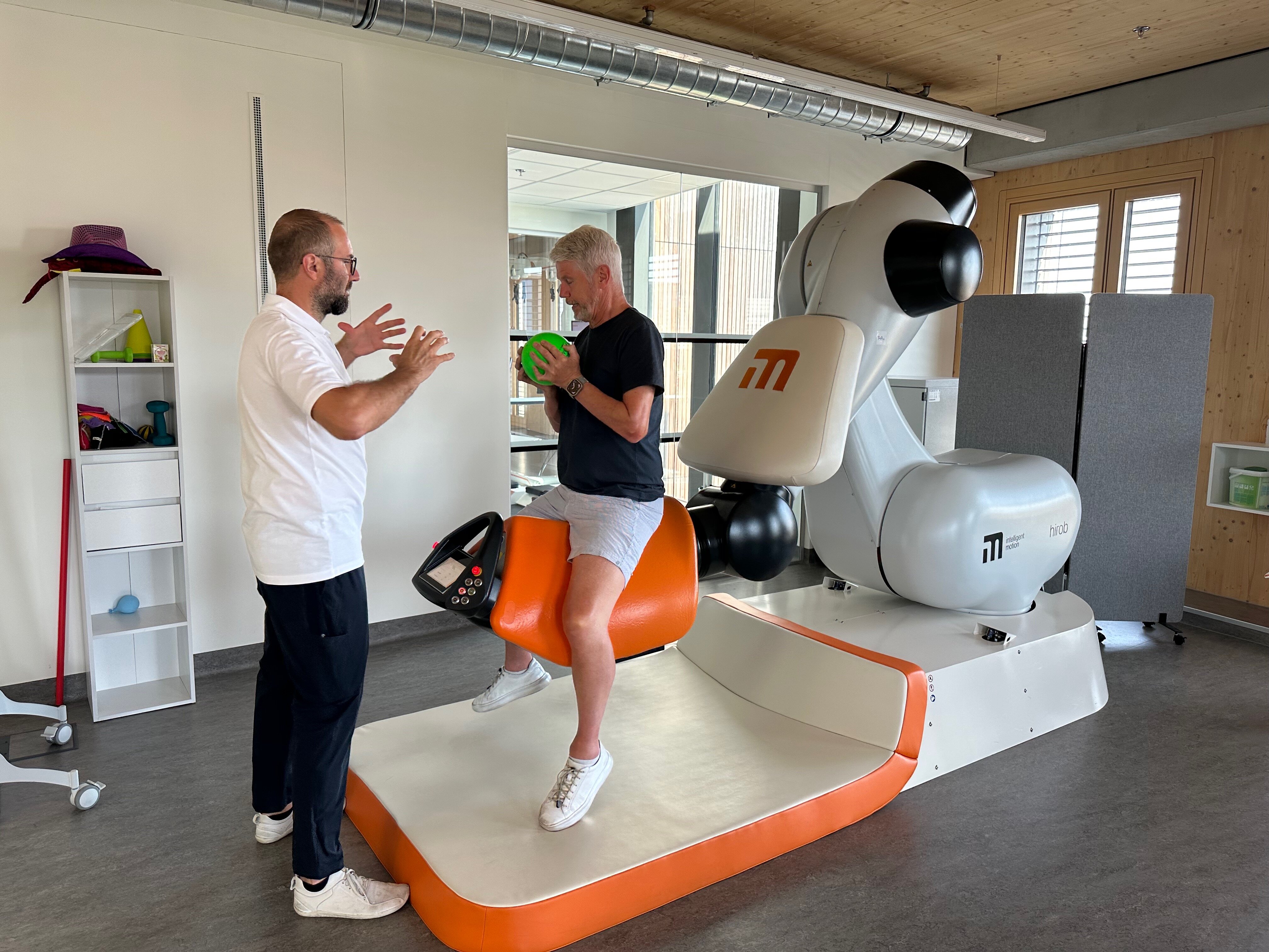 Ein Patient sitzt auf einer Liege mit einem Ball in der ausgestreckten Hand, der Therapeut steht vor ihm und zeigt ihm die Übung vor