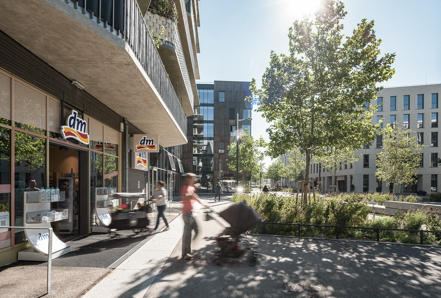 Auf der linken Seite eine Hausfassade mit einer Dm-Filiale, in der Mitte eine Person die einen Kinderwagen schiebt, diese ist verschwommen, auf der rechten Seite ein Platz mit einem begrünter Teil mit Baum, im Hintergrund Häuser