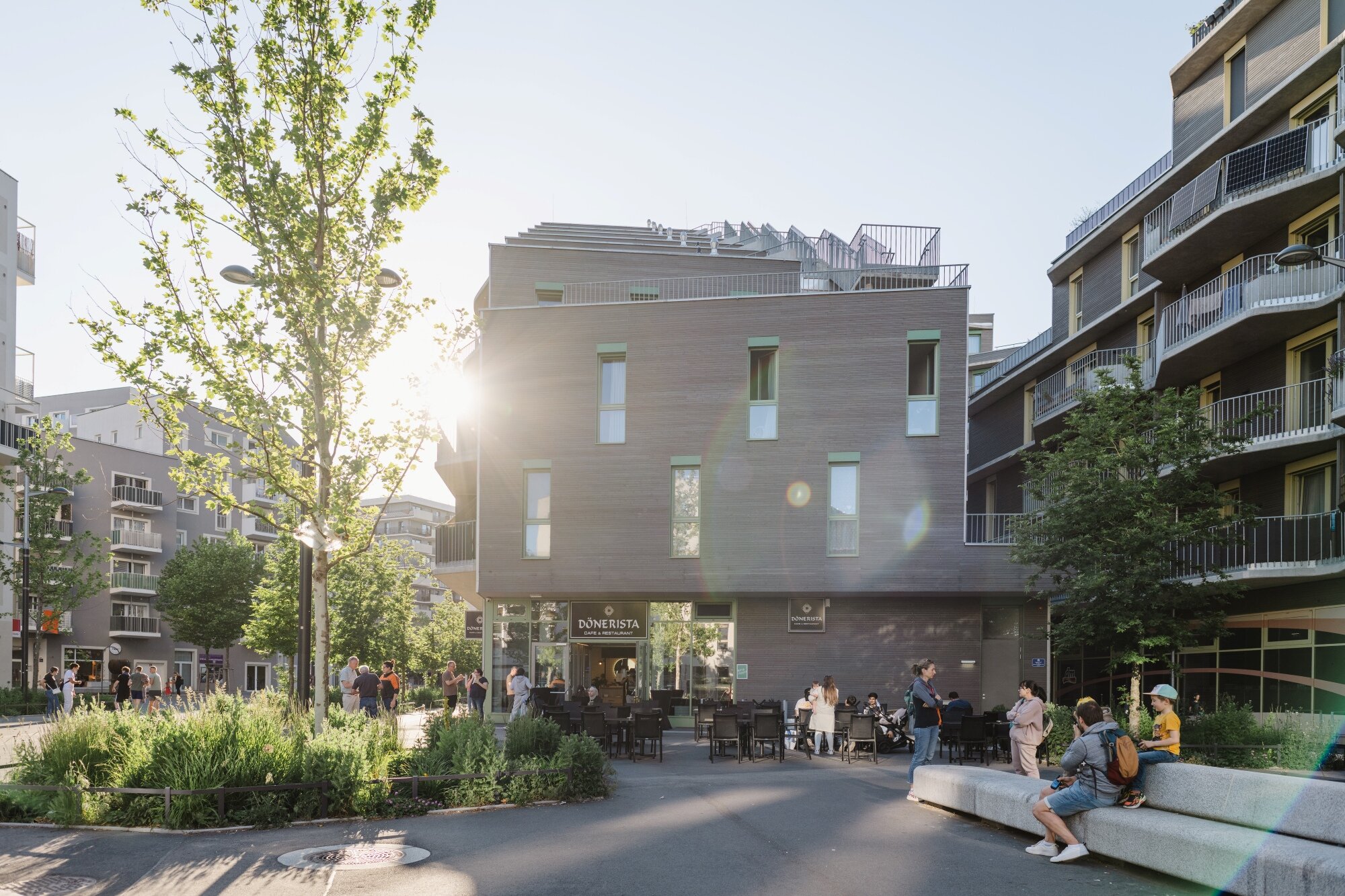 Menschen spatzieren und sitzen auf einem begrünten Platz mit Gastronomie und Shops
