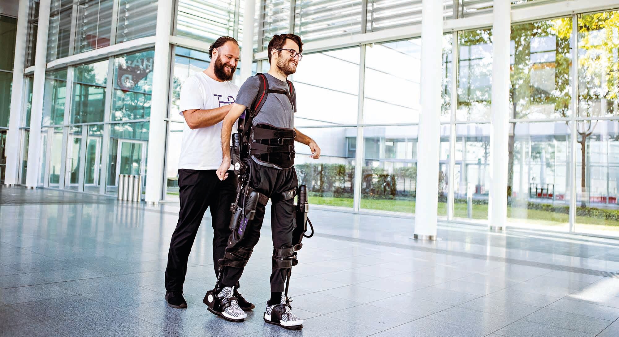 ein Mann begleitet einen anderen mit Exoskelett in einem modernen Gebäude beim Gehen