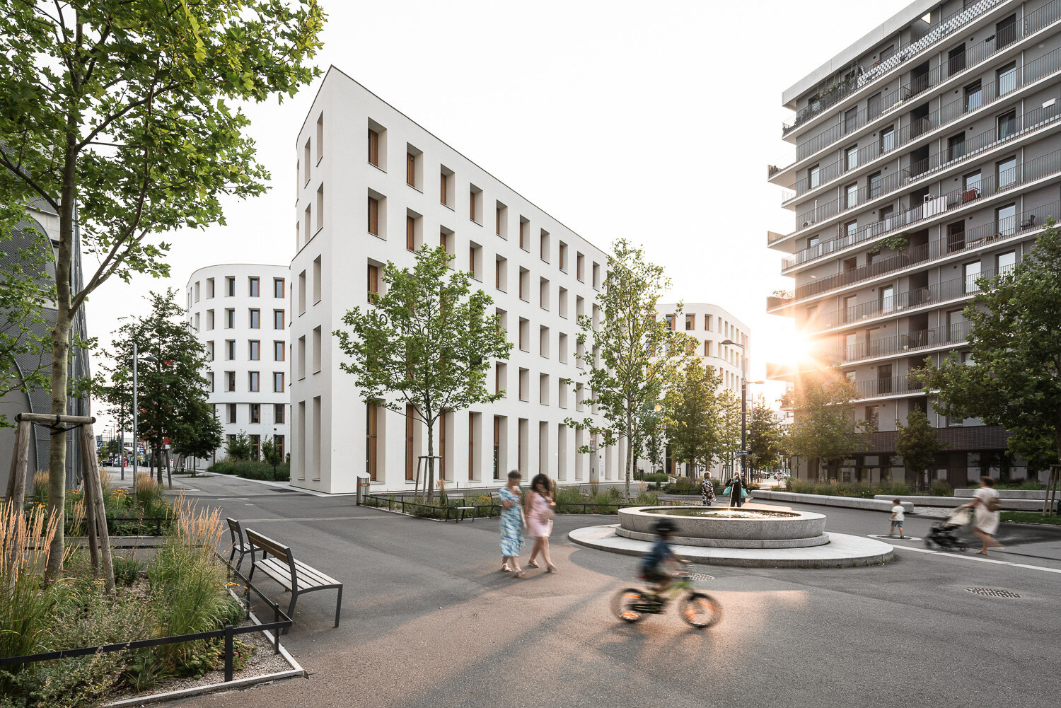 Zwei Frauen und Kinder auf einem Platz mit Bäumen und Brunnen und modernen Gebäude