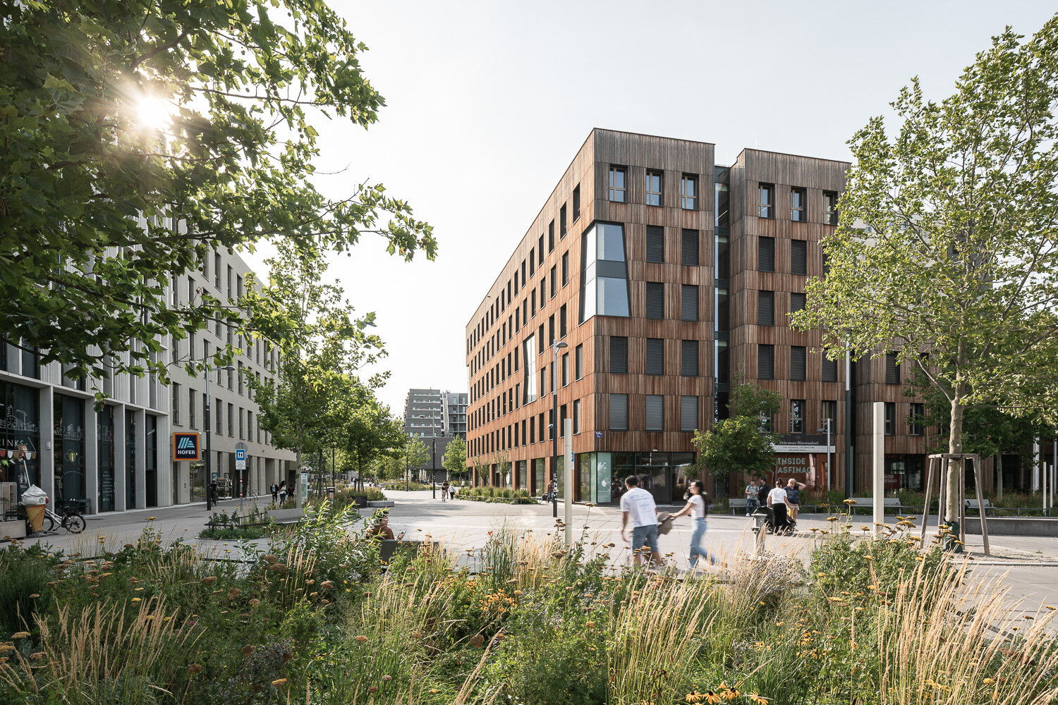 Menschen in einer begrünten Fußgängerzone, einem modernen Holzgebäude, Café und Shops