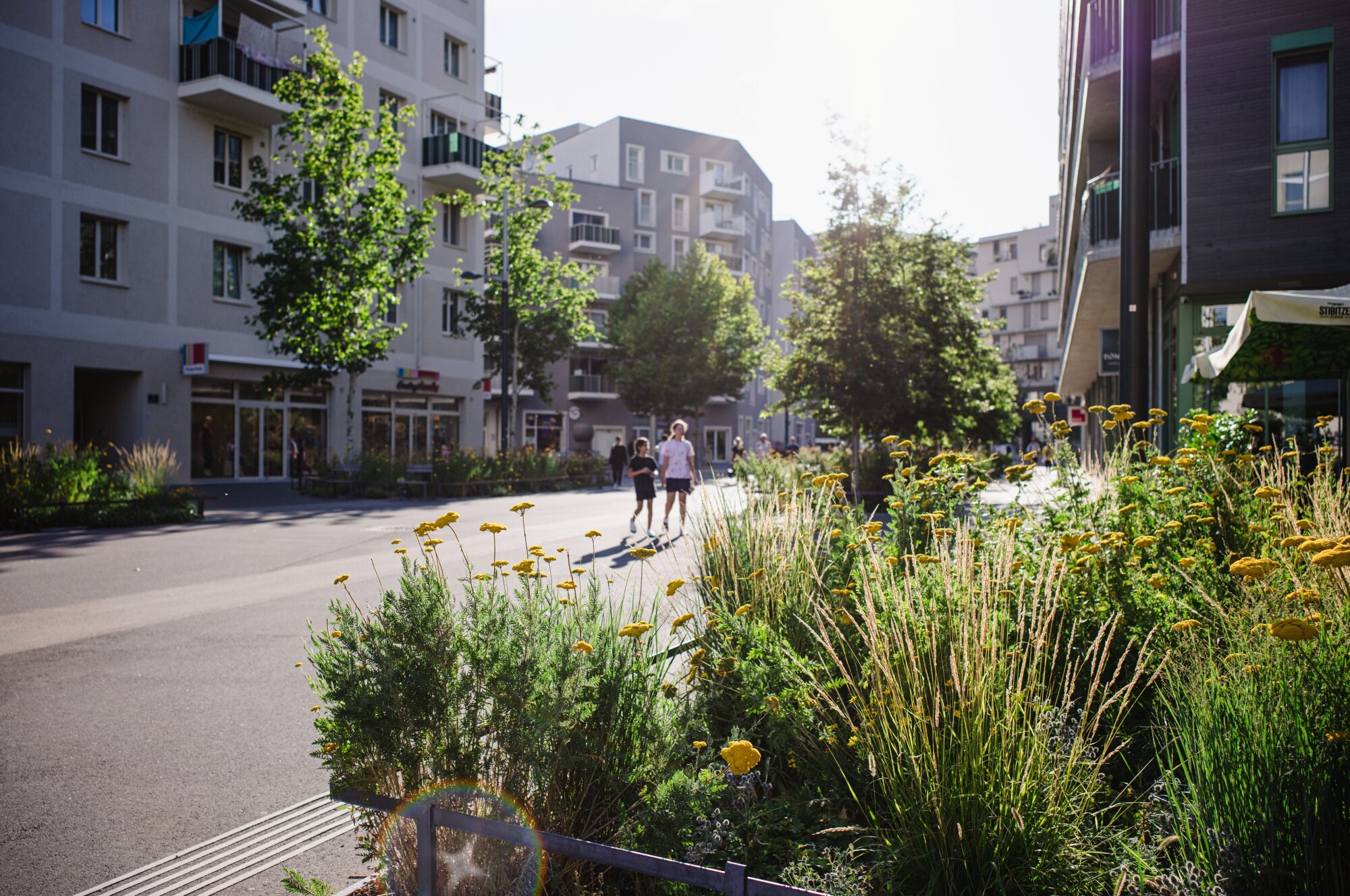 Menschen spazieren in einer begrünten Fußgängerzone zwischen Lokalen und Shops