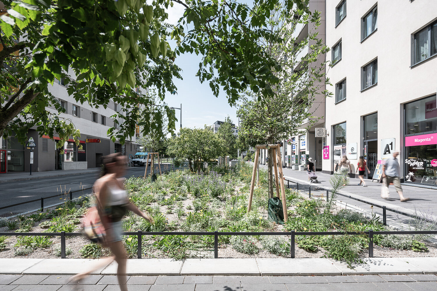 Frau geht entlang einer Einkaufsstraße mit mehreren Geschäften und viel Grün