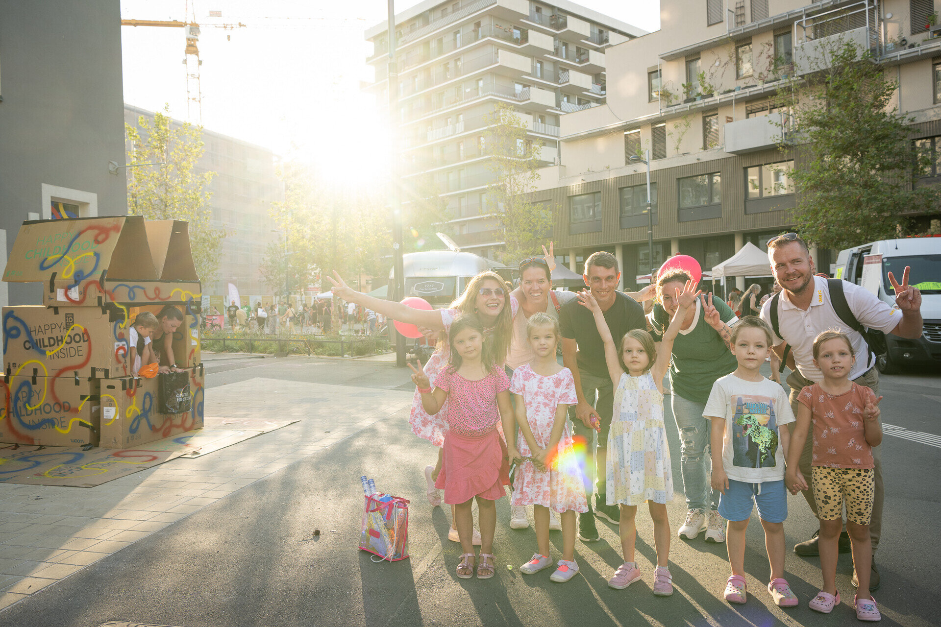 Gruppe von Kindern und Erwachsenen posiert lachend bei Sonnenschein auf einem Straßenfest in der Seestadt. Im Hintergrund sind moderne Wohnhäuser, ein bemaltes Papphaus und weitere Besucherinnen zu sehen.