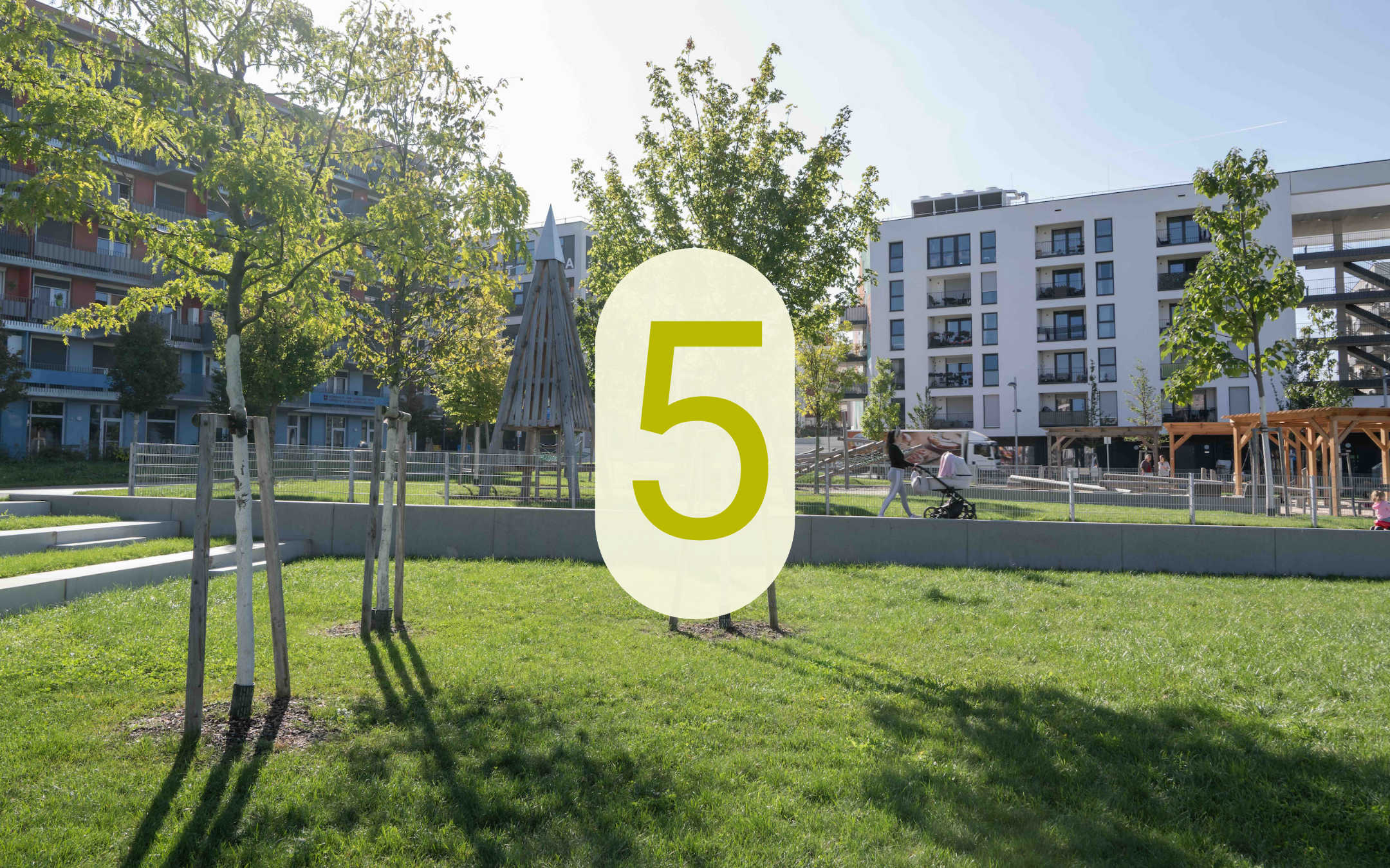 Grüne Freifläche in der Seestadt mit Bäumen, Spielplatz und Wohngebäuden sowie der eingeblendeten Zahl 5.