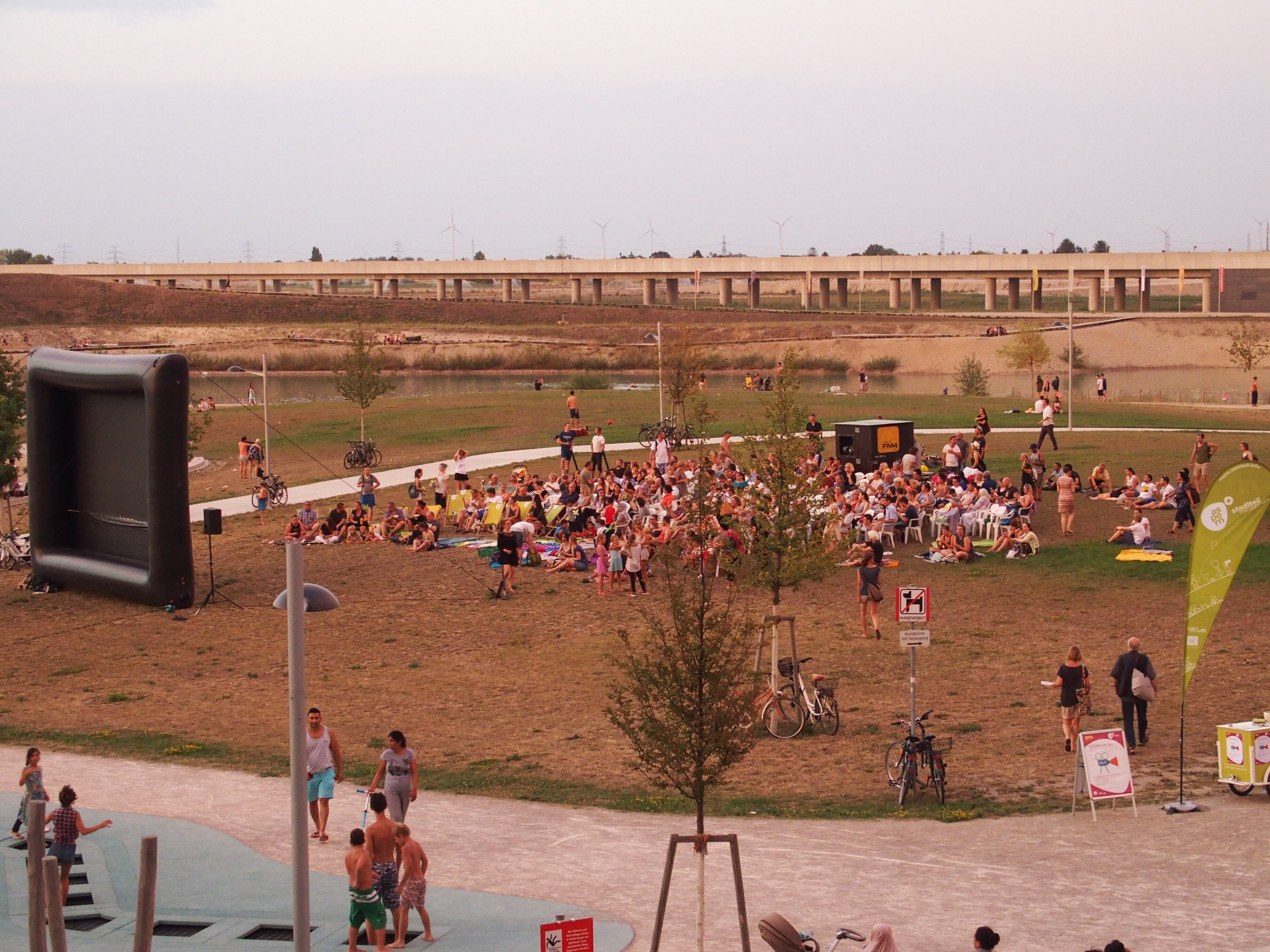 Menschen im Seepark, die vor der Filmleinwand im Gras sitzen. Im Hintergrund ist der See und die Ubahn zu sehen. Fotografiert von oben.