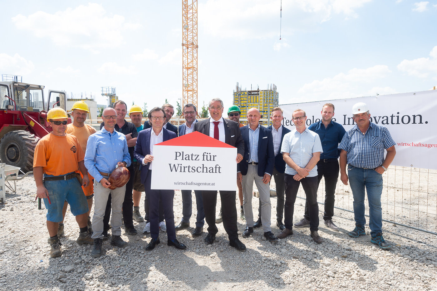 Mehrere Personen mit einem Schild mit der Aufschrift "Platz für Wirtschaft". Im Hintergrund Baustelle. 