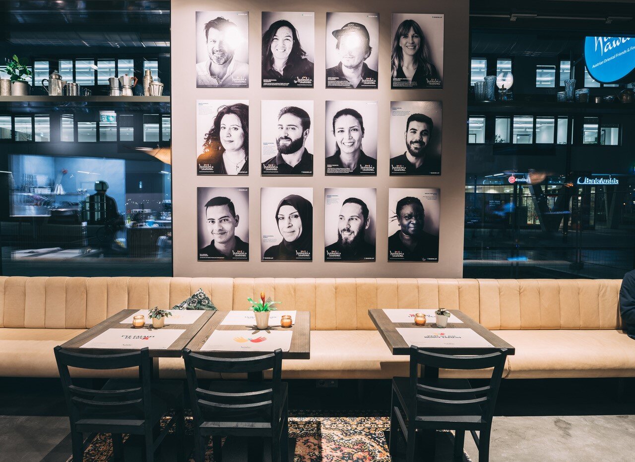 Innenansicht eines Lokals mit Tischen, Sesseln. Im Hintergrund Porträts von Menschen. 