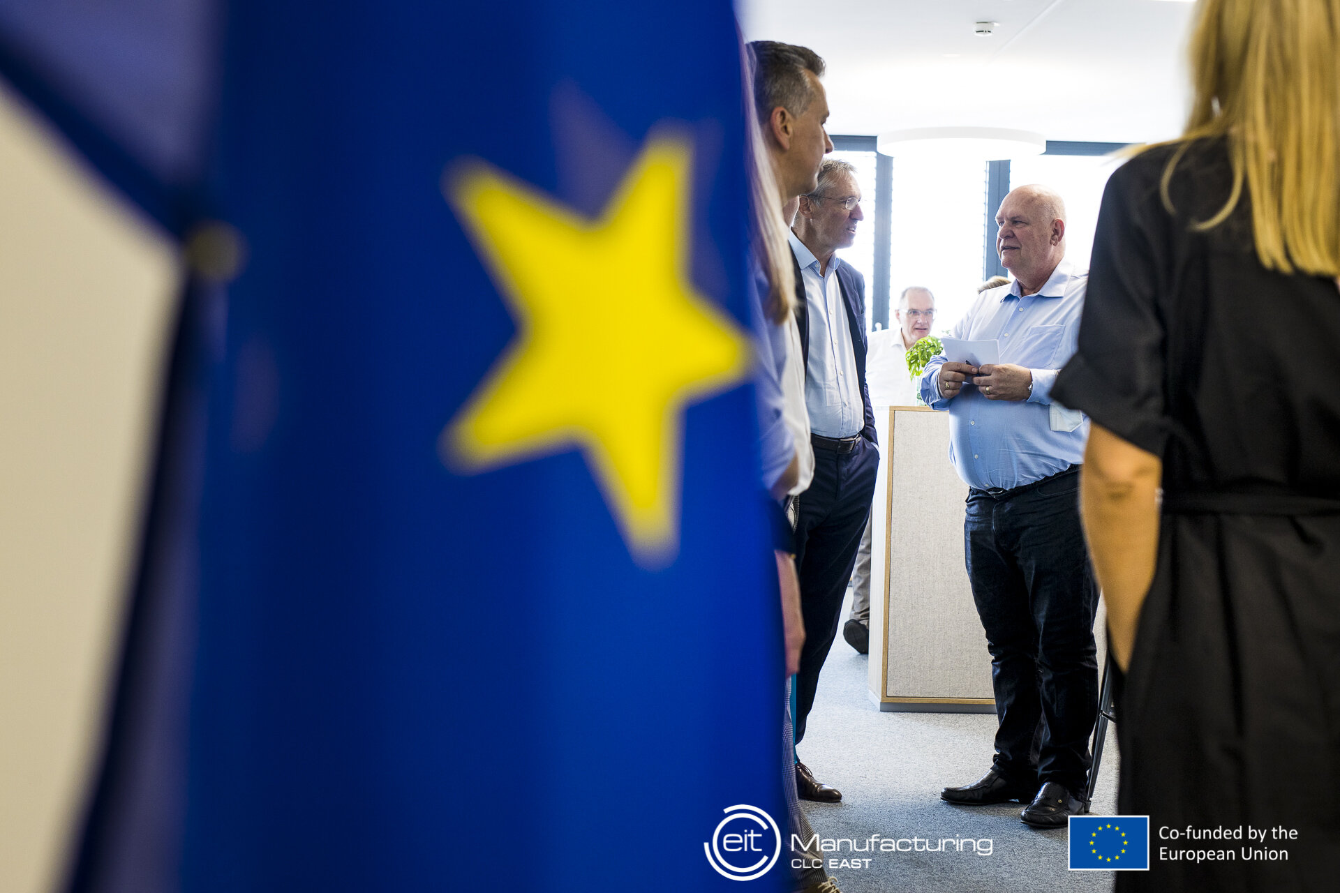 EU-Fahne im Vordergrund, dahinter sprechende Leute bei der Veranstaltung. 