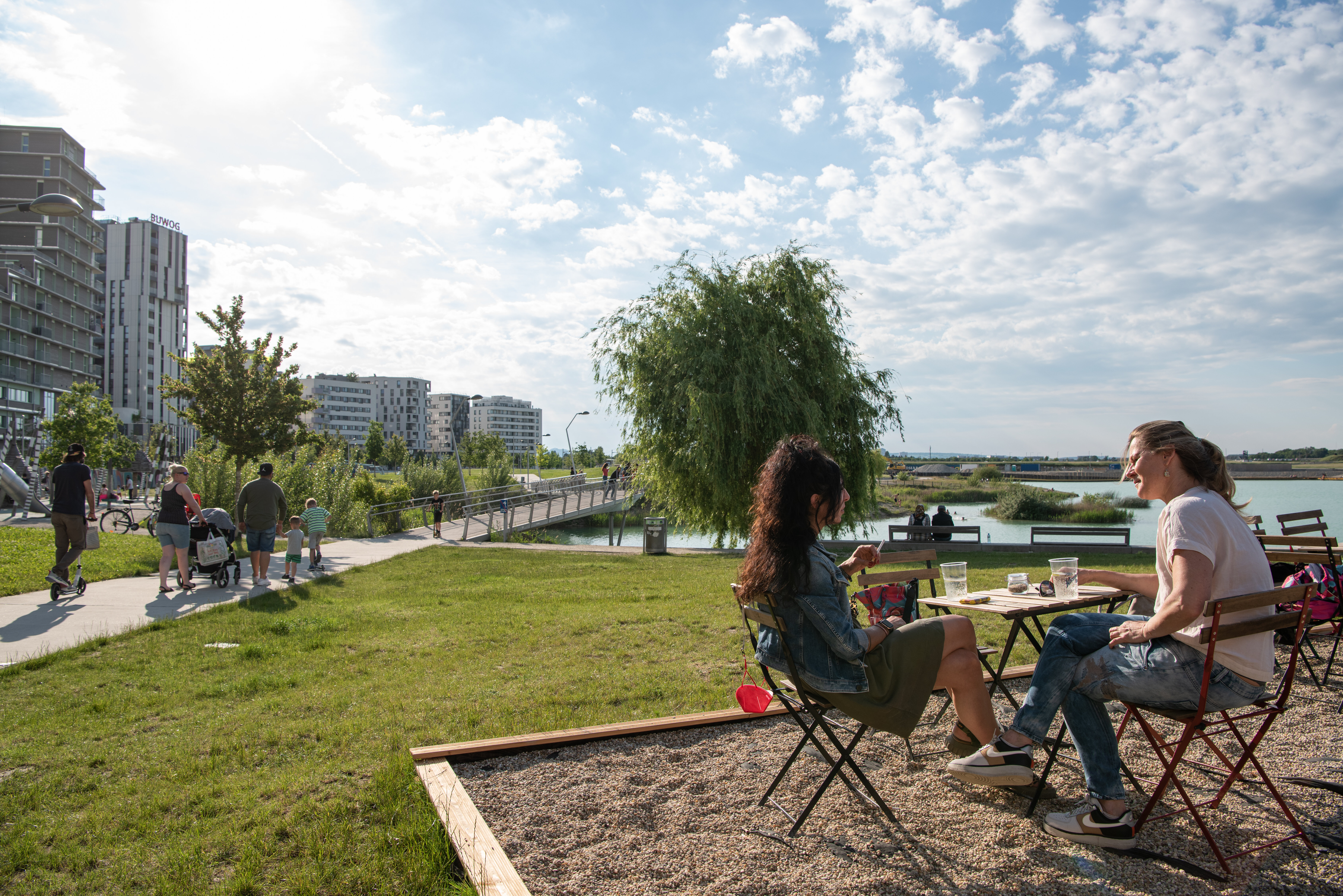 Zwei Personen in einer Outdoor-Bar, im Hintergrund Seepark mit Spaziergängern. 