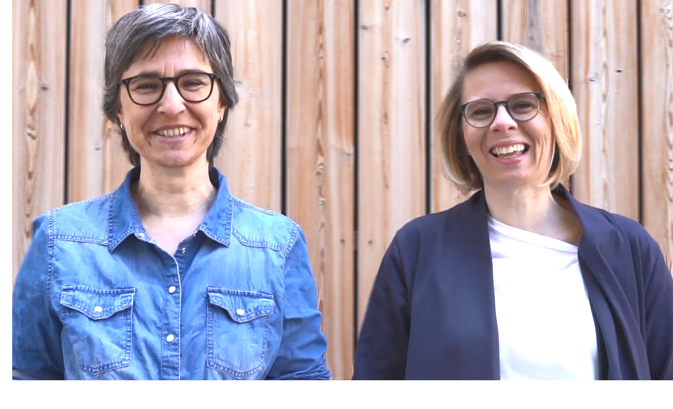 Martina und Eva-Maria Halmer stehen lächelnd vor einer Holzwand.
