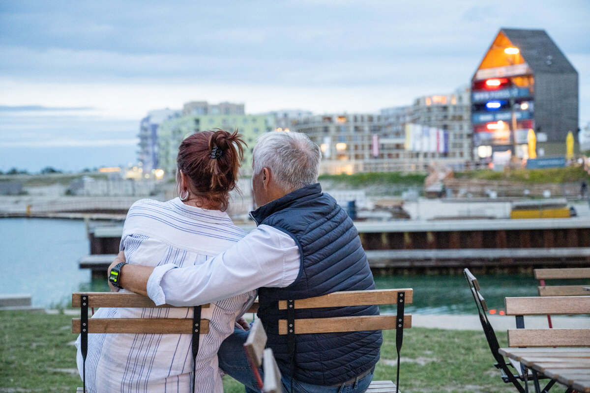 Zwei Personen männlich und weiblich auf einer Bank in der Seestadt - von hinten in die Ferne blickend. See im Hintergrund. 
