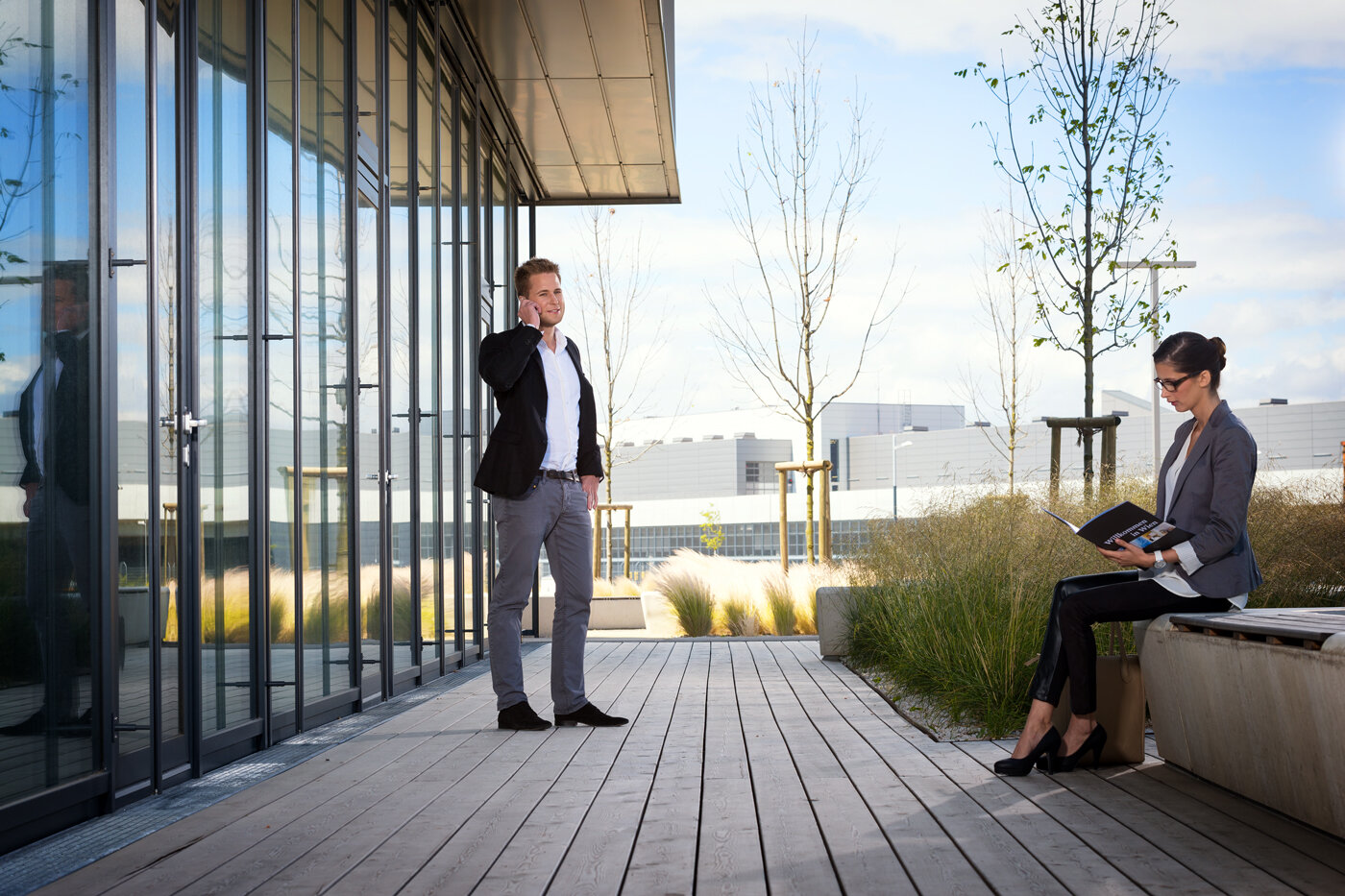 Terrasse: Ein Mann telefoniert, eine Frau sitzt und liest in einer Zeitschrift. Businesslook. Links moderne Glasfassade. 