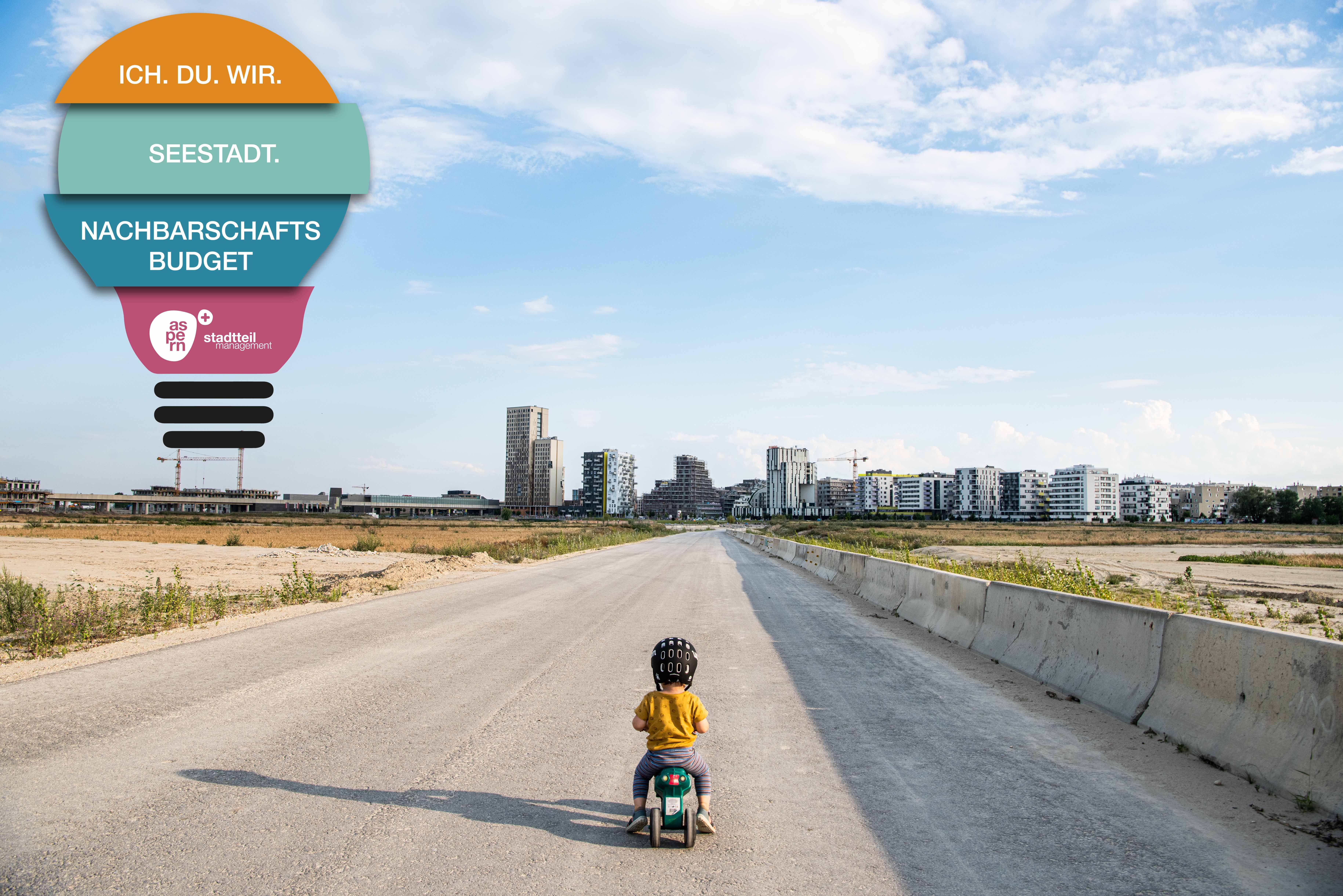 Ein kleines Kind fährt mit Kinderspielfahrzeug auf einer Straße Richtung Seestadt.