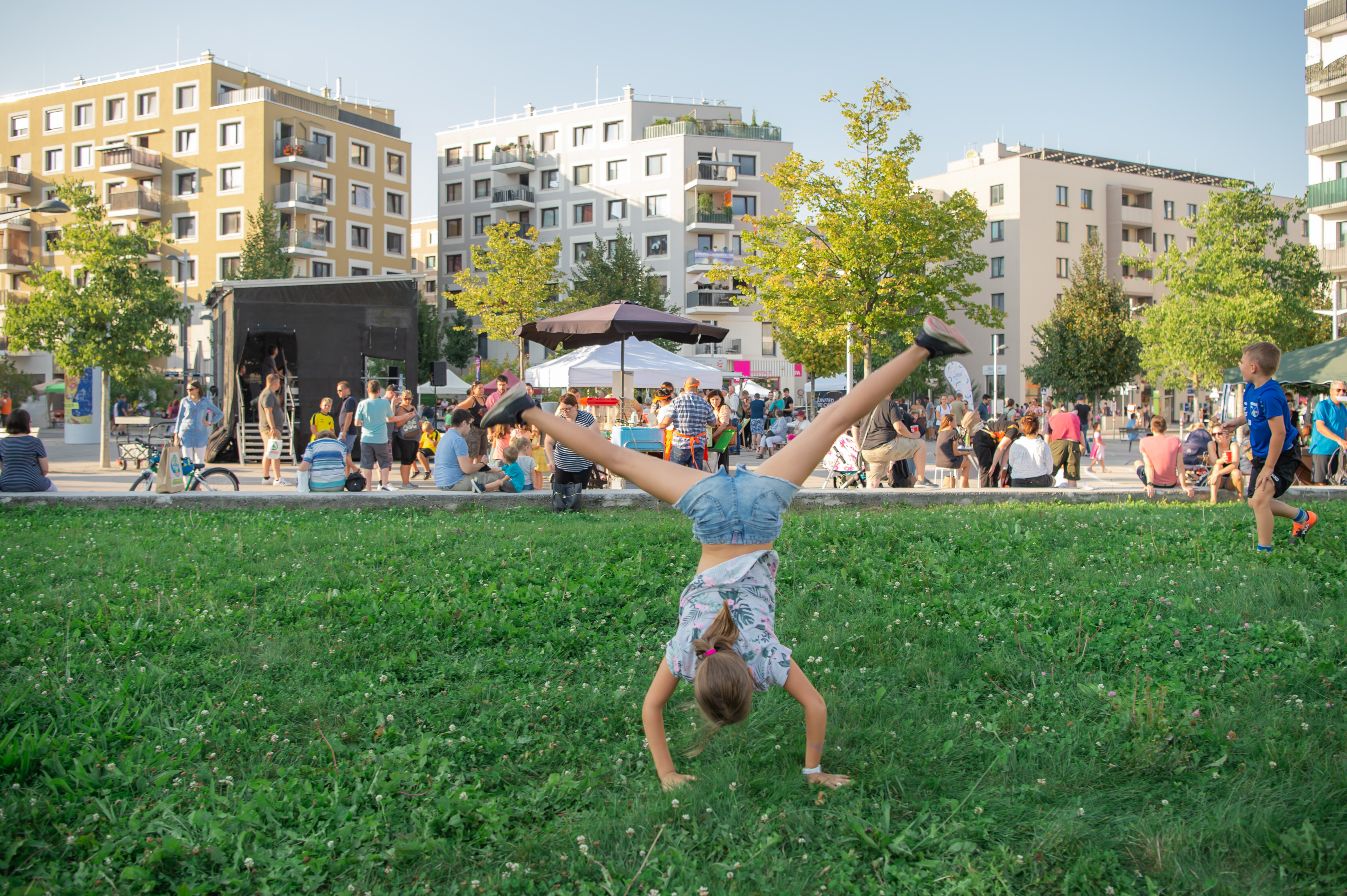 Ein Kind schlägt ein Rad auf einer Wiese. Dahinter findet ein Nachbarschaftsfest statt.
