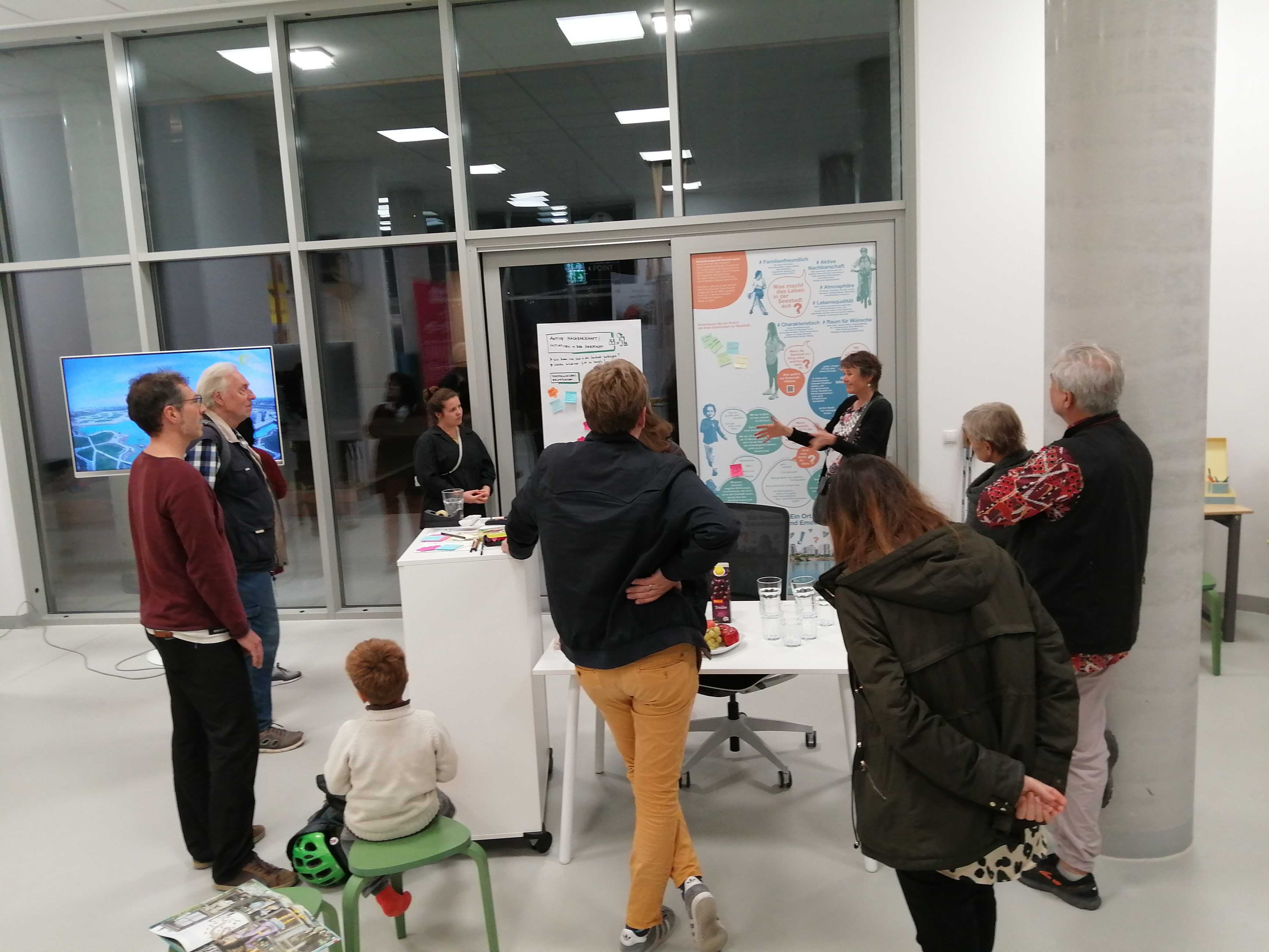 Eine Gruppe von Menschen steht in einem Raum vor einem Poster zum Thema "So lebt es sich in der Seestadt". 