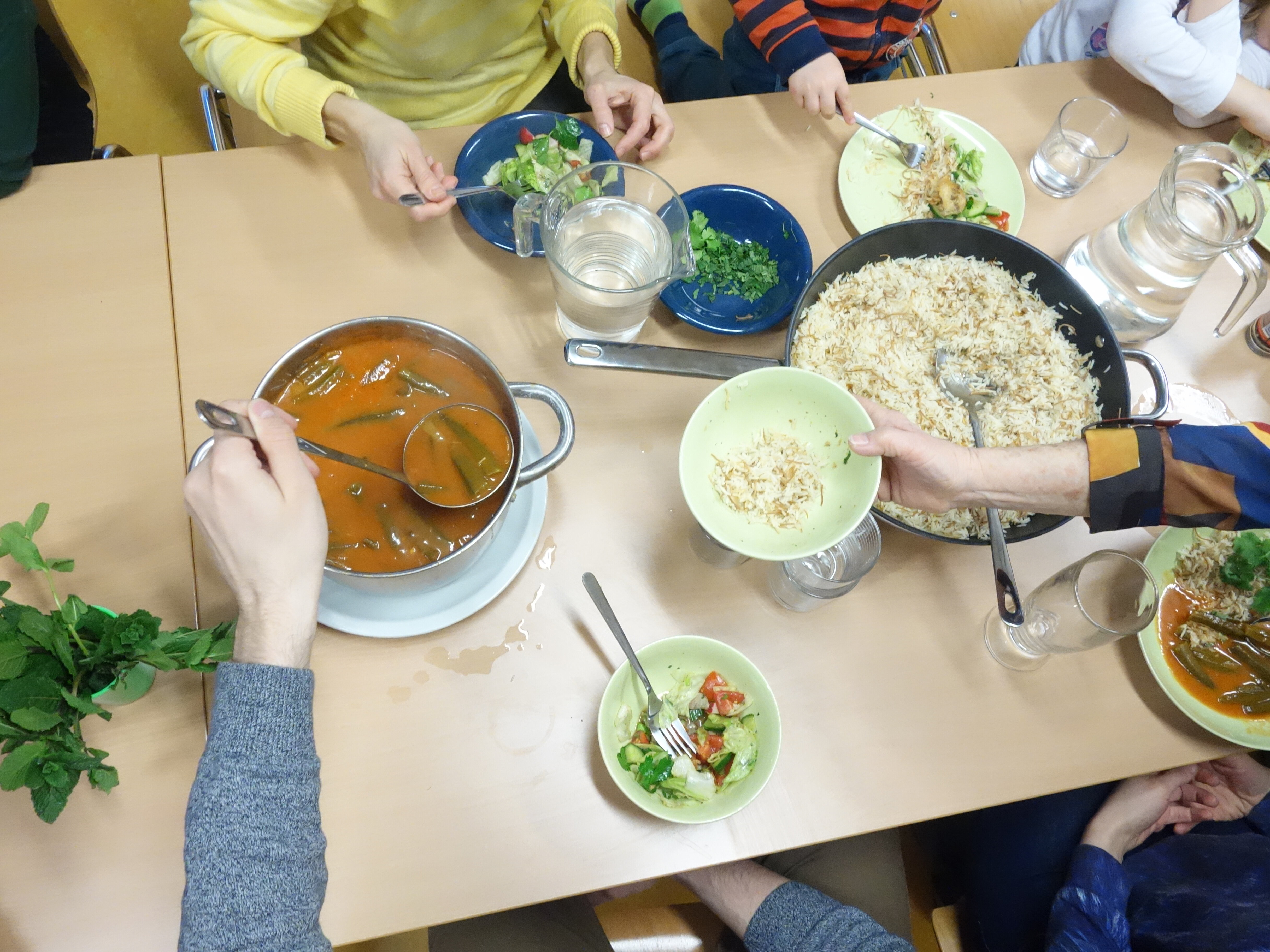 Auf einem Tisch stehen ein Eintopf und eine Pfanne Reis. Man sieht Hände von Menschen, die sich gerade Essen auf Teller geben.