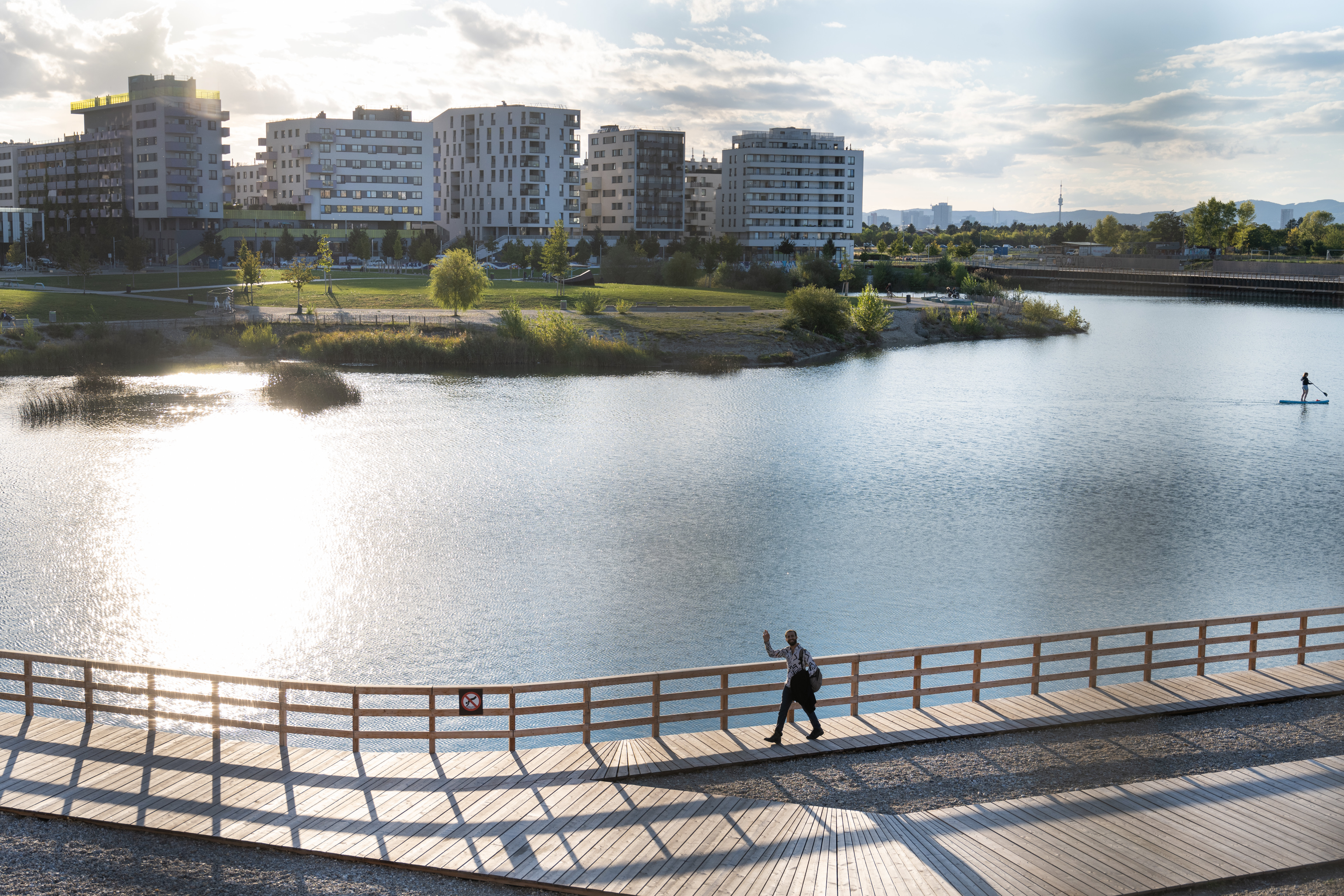 Ein Mann geht am Steg entlag des Seestadt Sees und winkt dem/der Betrachter*in zu. Im Hintergrund sieht man Häuser der Seestadt.