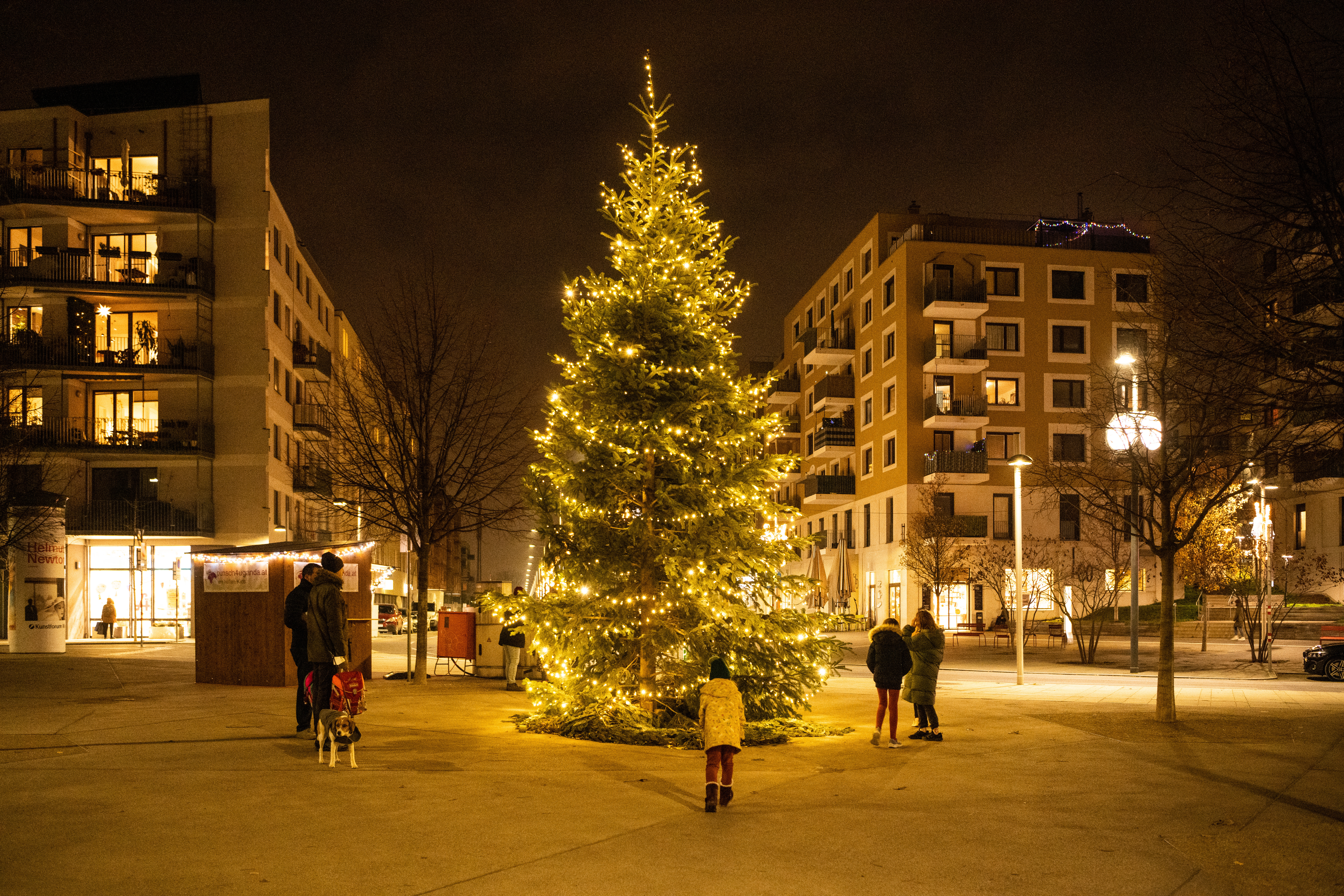 Rundum einen großen beleuchteten Christbaum sammeln sich Kinder und Erwachsene. Im Hintergrund sieht man Häuser und einen Punschstand. 
