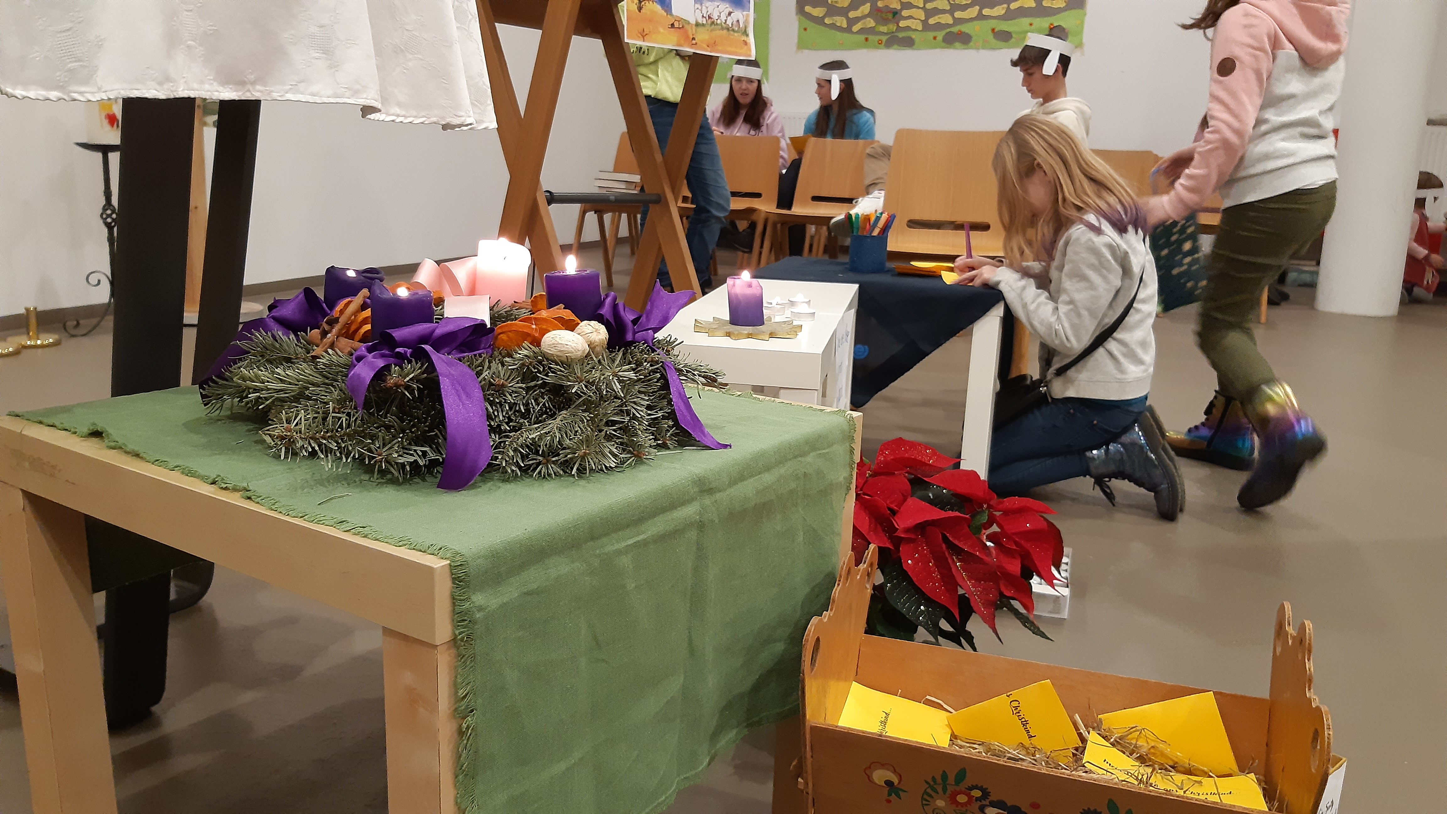 Im Vordergrund steht ein Adventkranz auf einem kleinen Tischchen, im Hintergrund basteln Kinder und Jugendliche