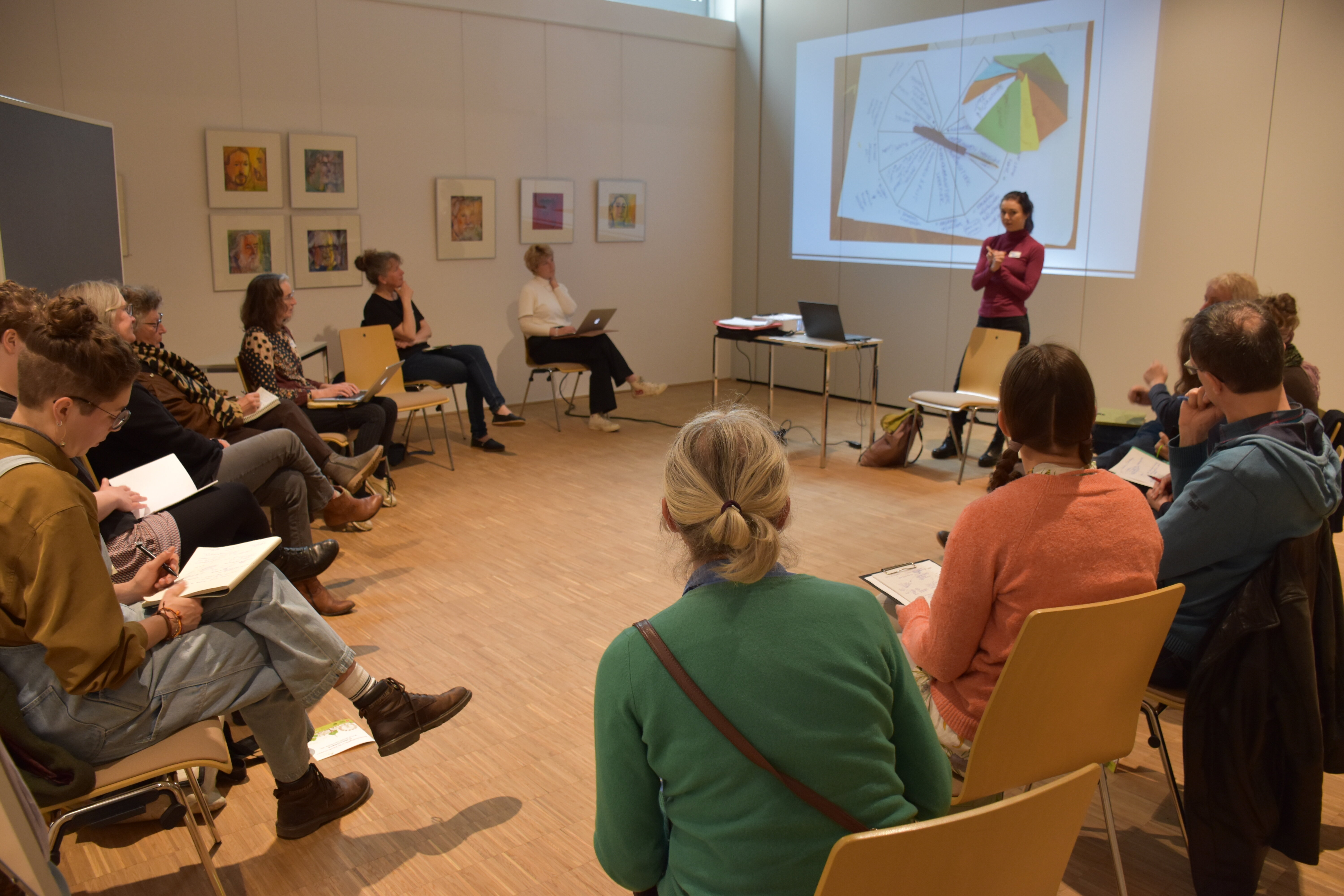 Mescnhen sitzen im Sesselkreis und schauen auf ein Präsentation auf der Wand