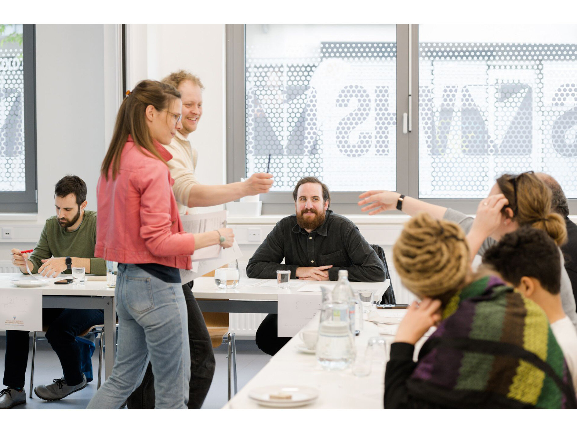 Eine Person reicht einer anderen Person einen Stift. Rundherum sitzen Menschen