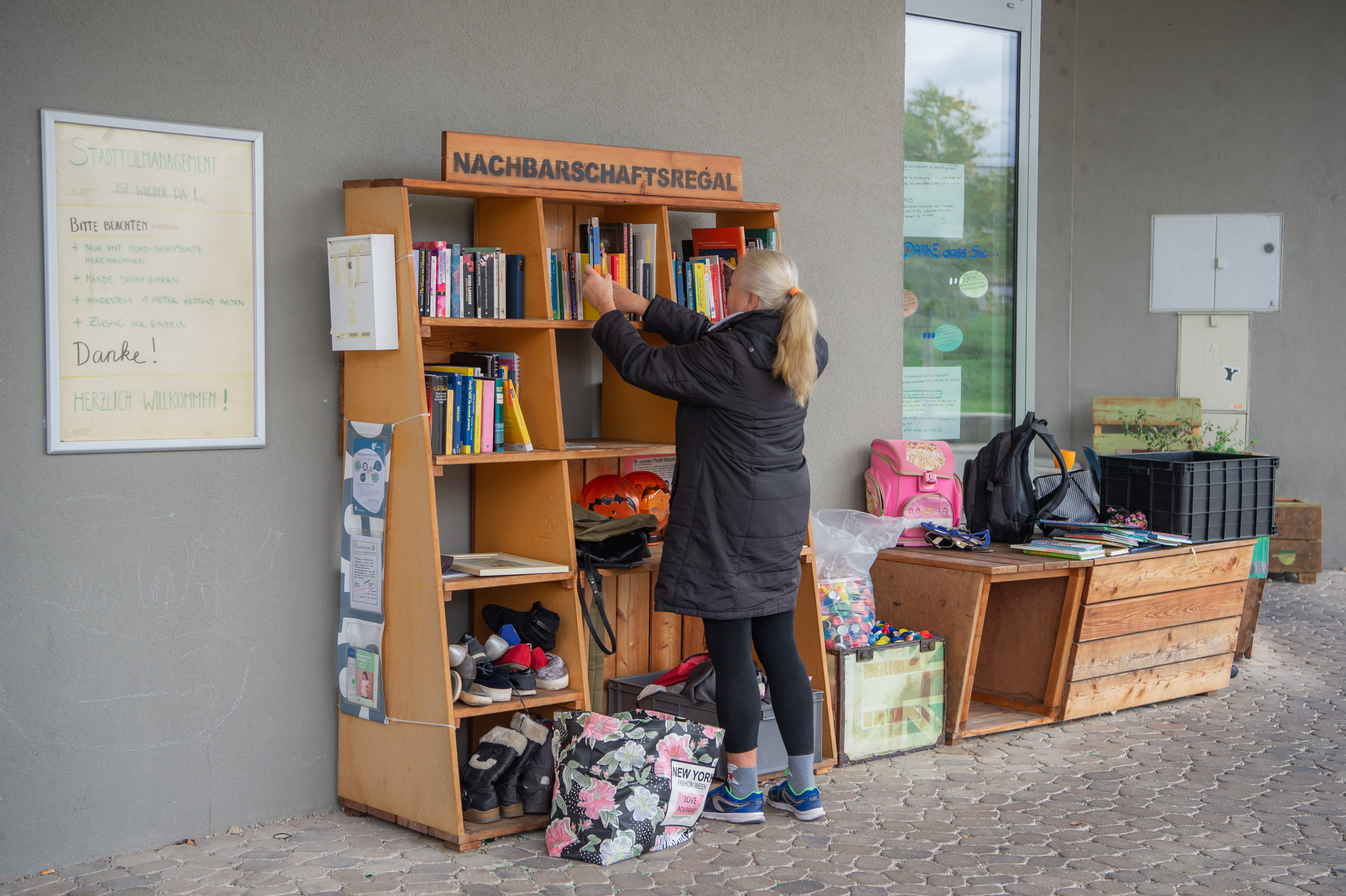 eine Frau mit Pferdeschwanzfrisur steht an einem Regal im Freien und nimmt sich was heraus
