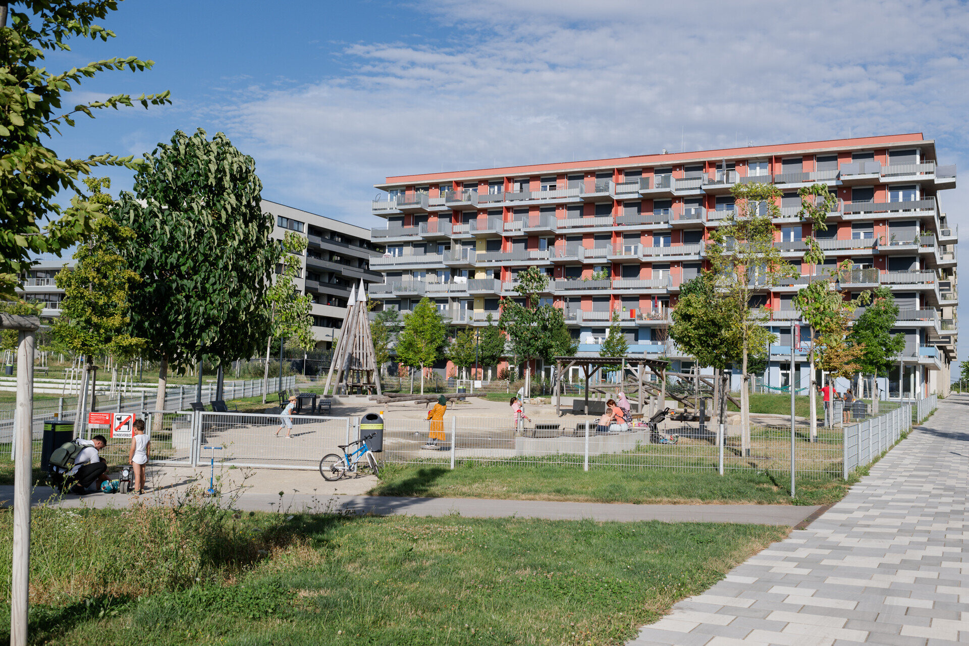 Ein farbenfrohes Wohngebäude, davor ein Spielplatz mit Kindern und Erwachsenen.