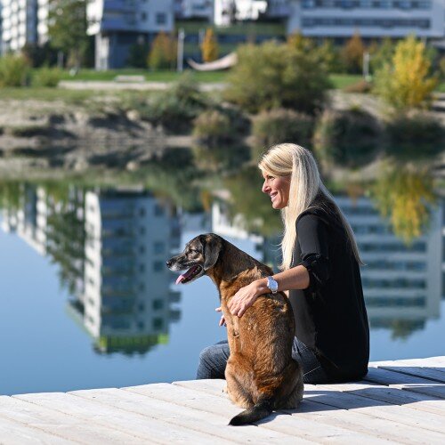 Tanja Bruny Eichinger am See sitzend. Hinter neben ihr ein Hund. Beide blicken in die Ferne.