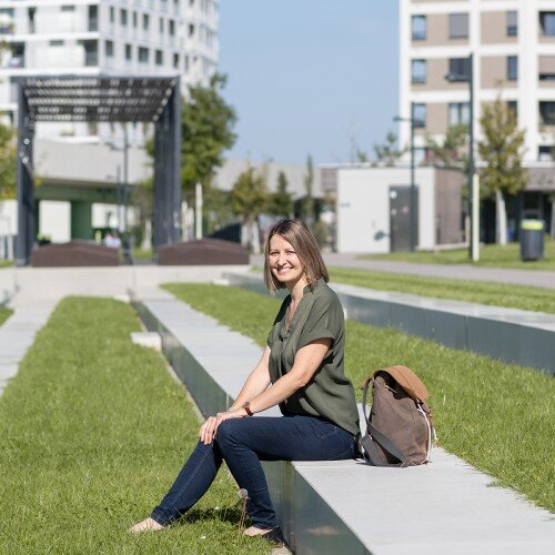 Annemarie Müller sitzend auf einer Bank auf einer Wiese. Hinter ihr sind Gebäude zu sehen.