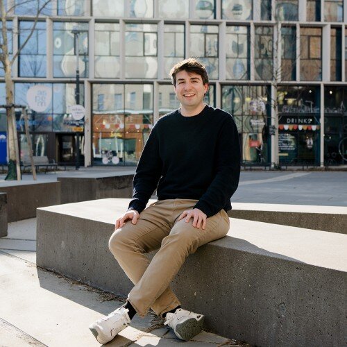 Sebastian Zenz sitzend auf einer modernen Bank. Hinter ihm ist ein großes, verglastes Gebäude.