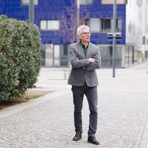 Harald Schlee spaziert über einen gepflasterten Weg. Hinter ihm ein blaues Gebäude.